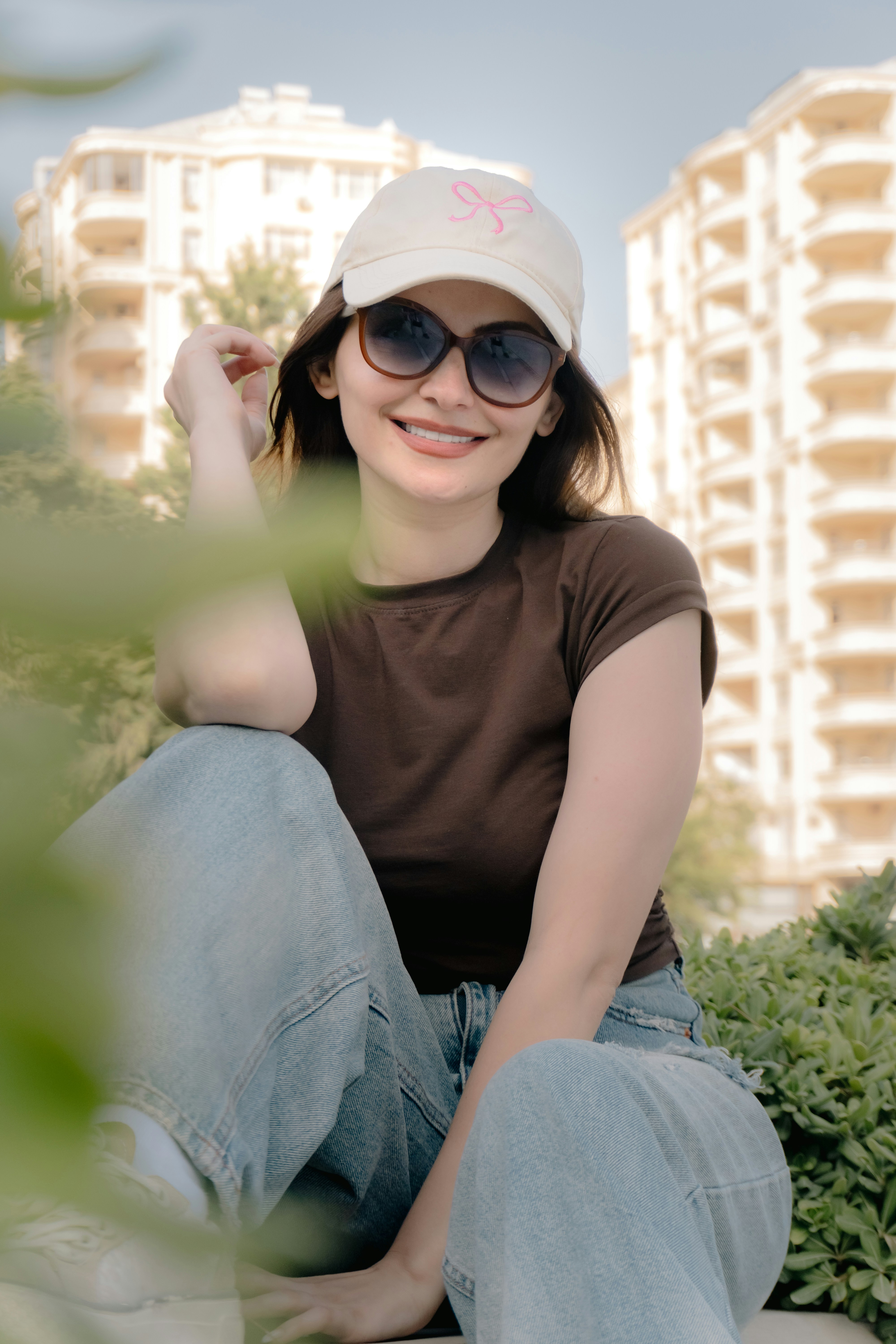 A woman in a brown t-shirt, jeans, and cap sitting outdoors near buildings