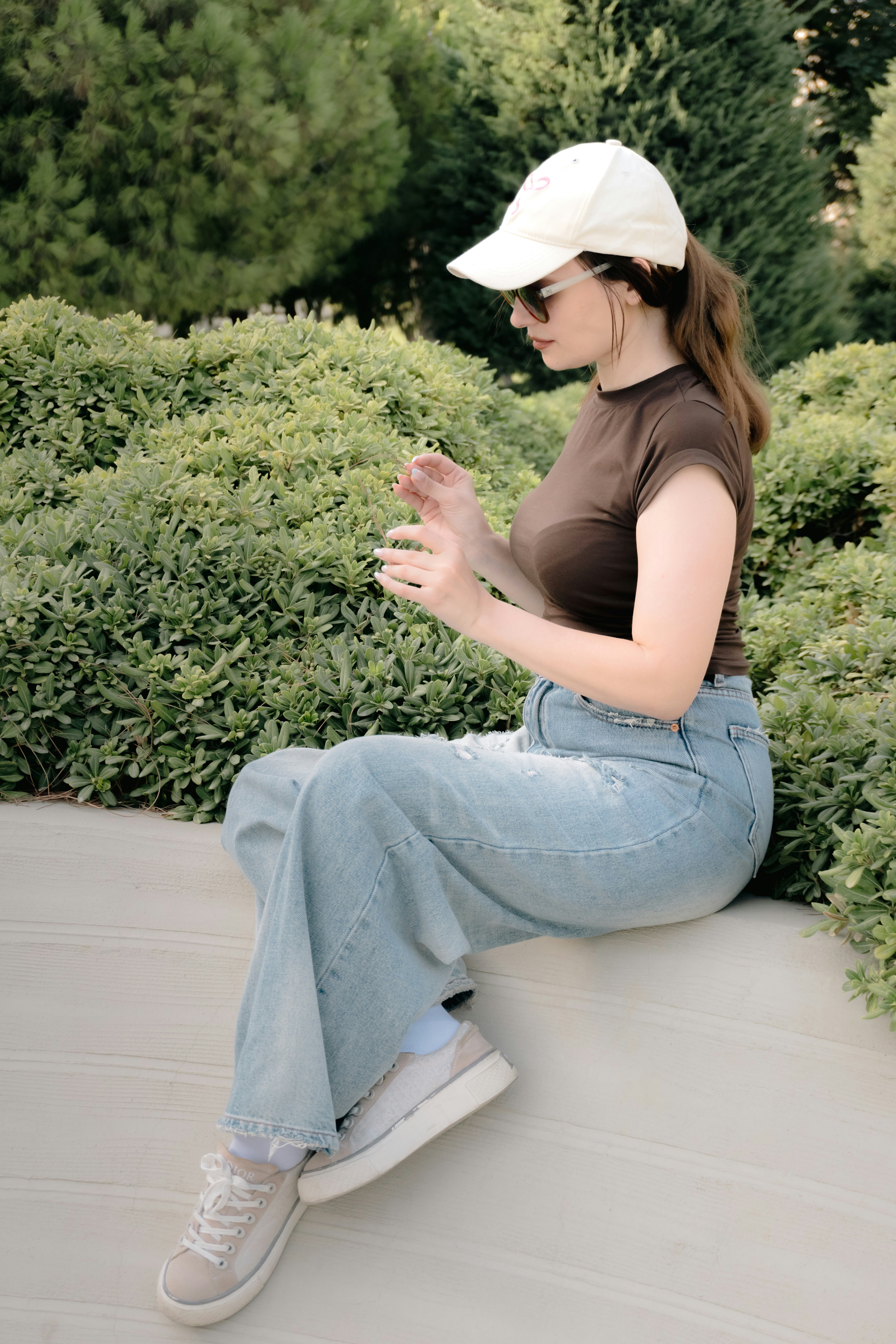 A woman in a brown t-shirt and jeans sitting on a ledge by bushes