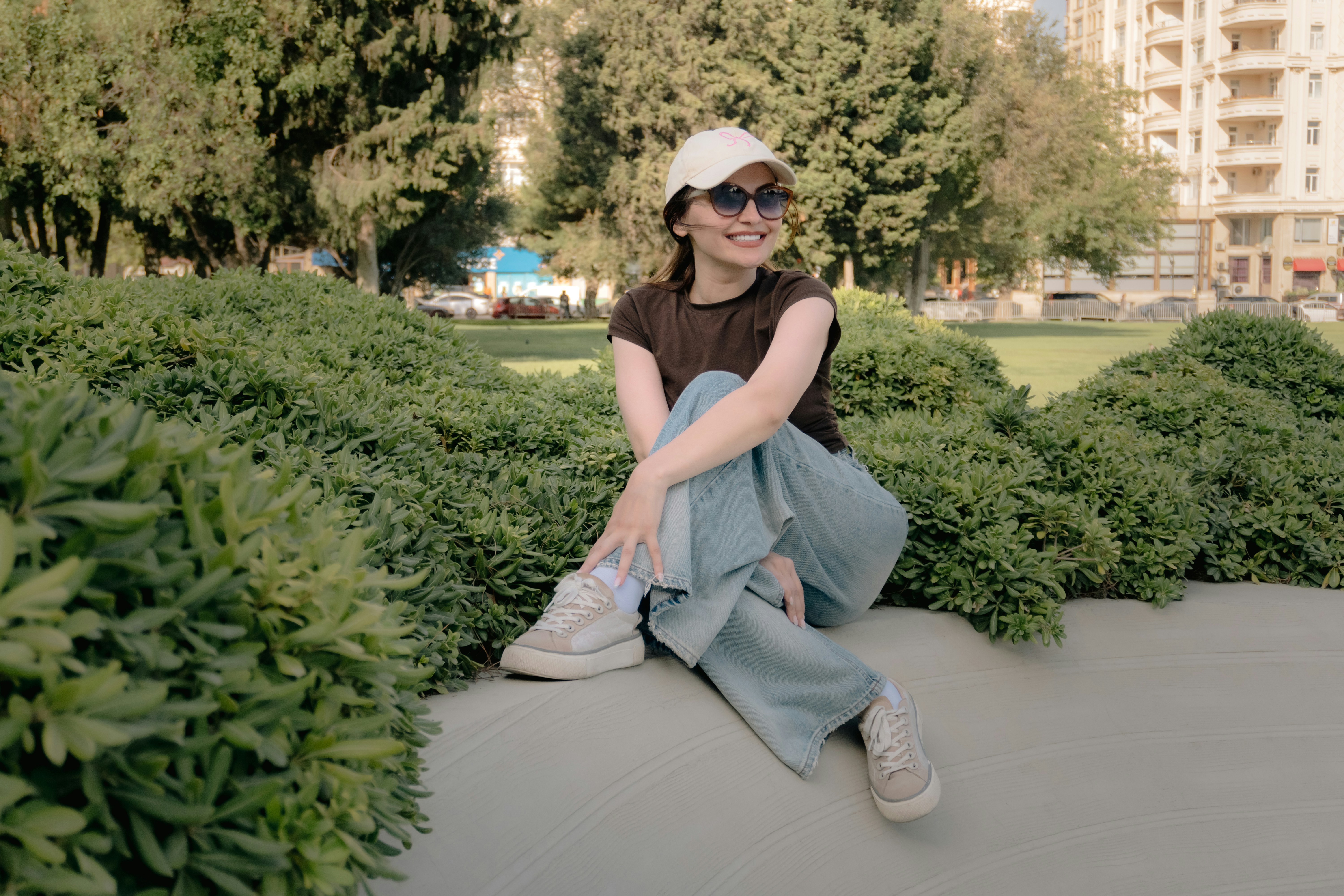 A woman in a brown t-shirt and jeans sitting among green bushes in a park