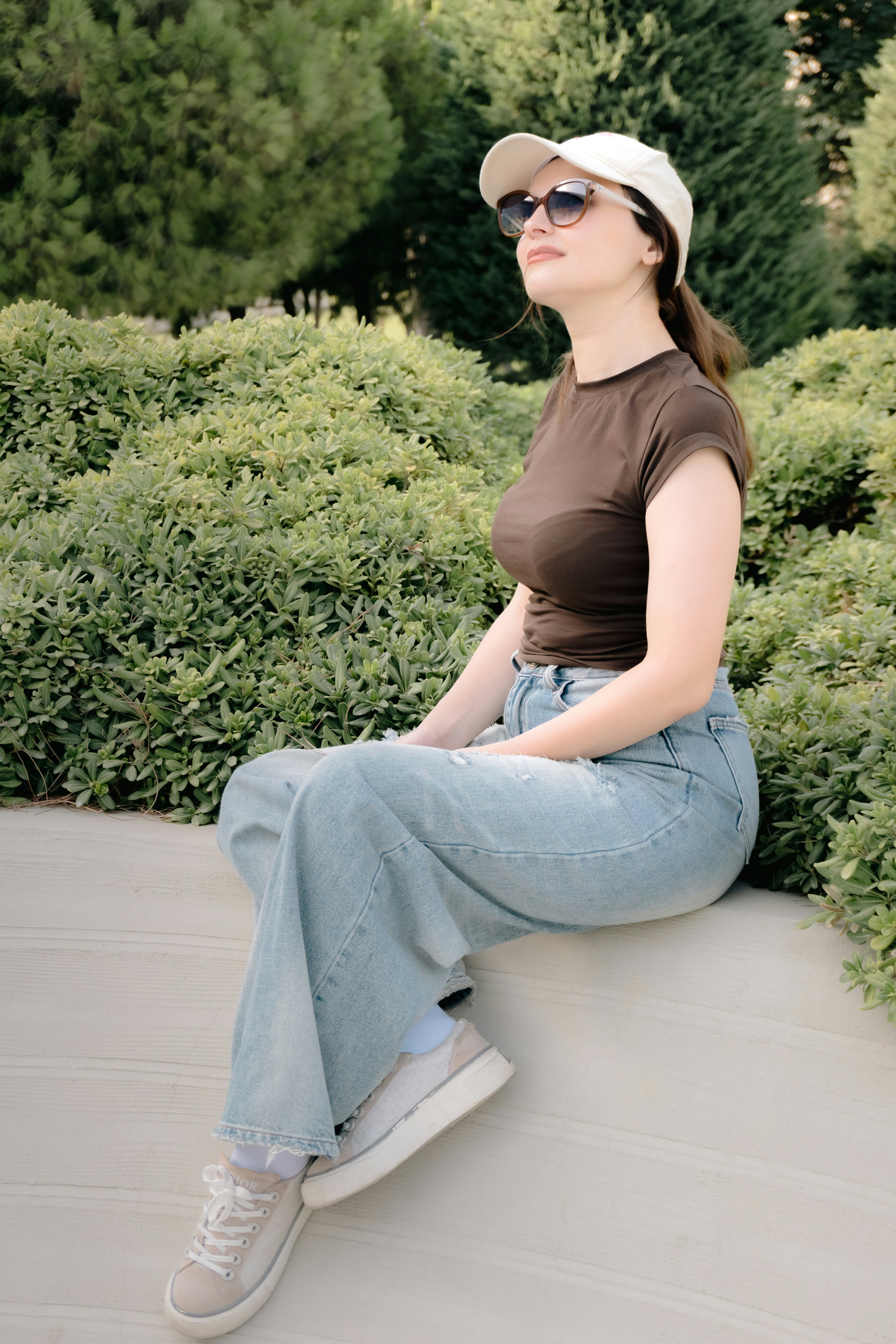 A woman in a brown t-shirt and jeans sitting on a ledge by bushes