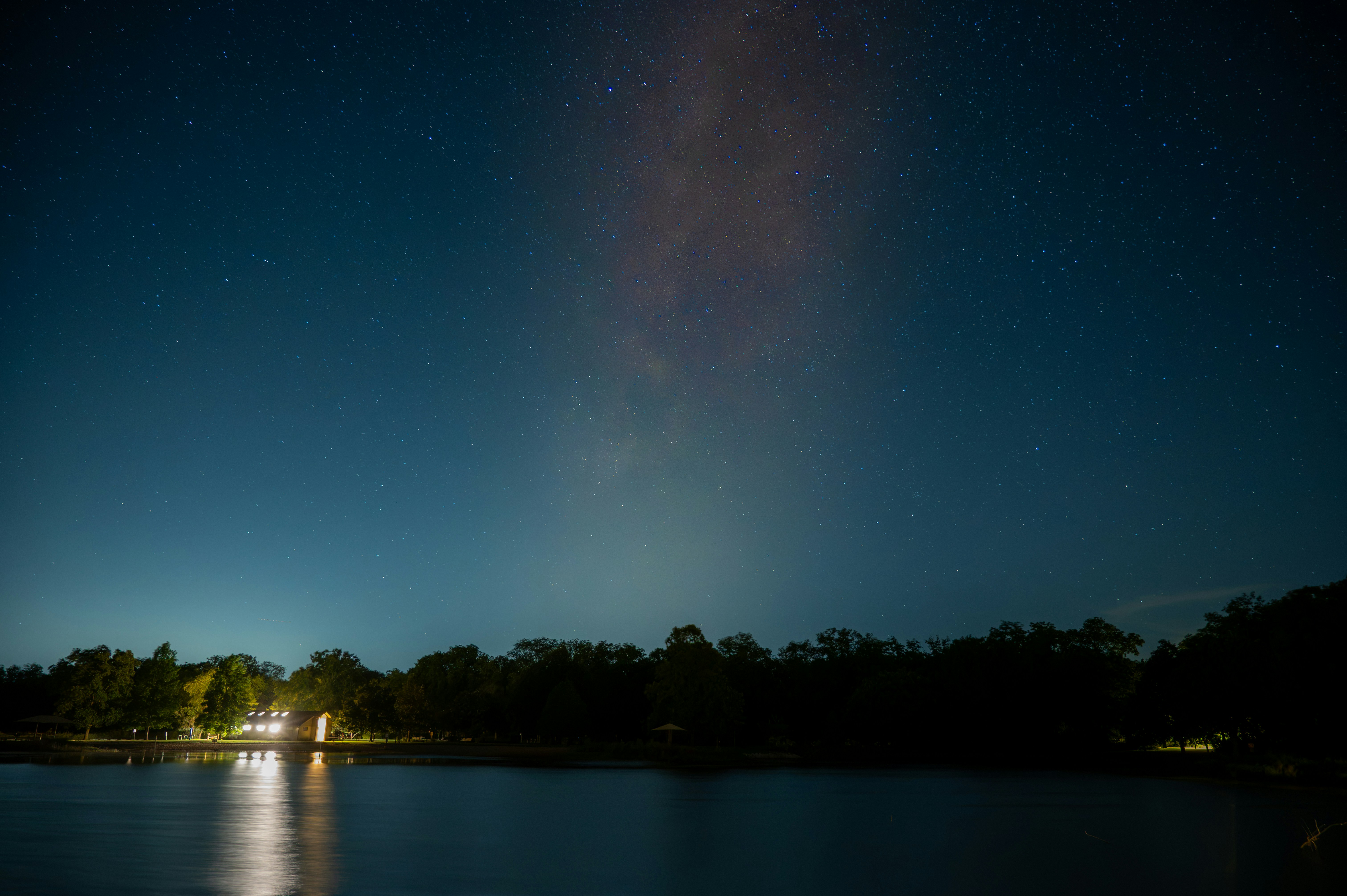 A bright building reflected in a dark lake under a starry night sky