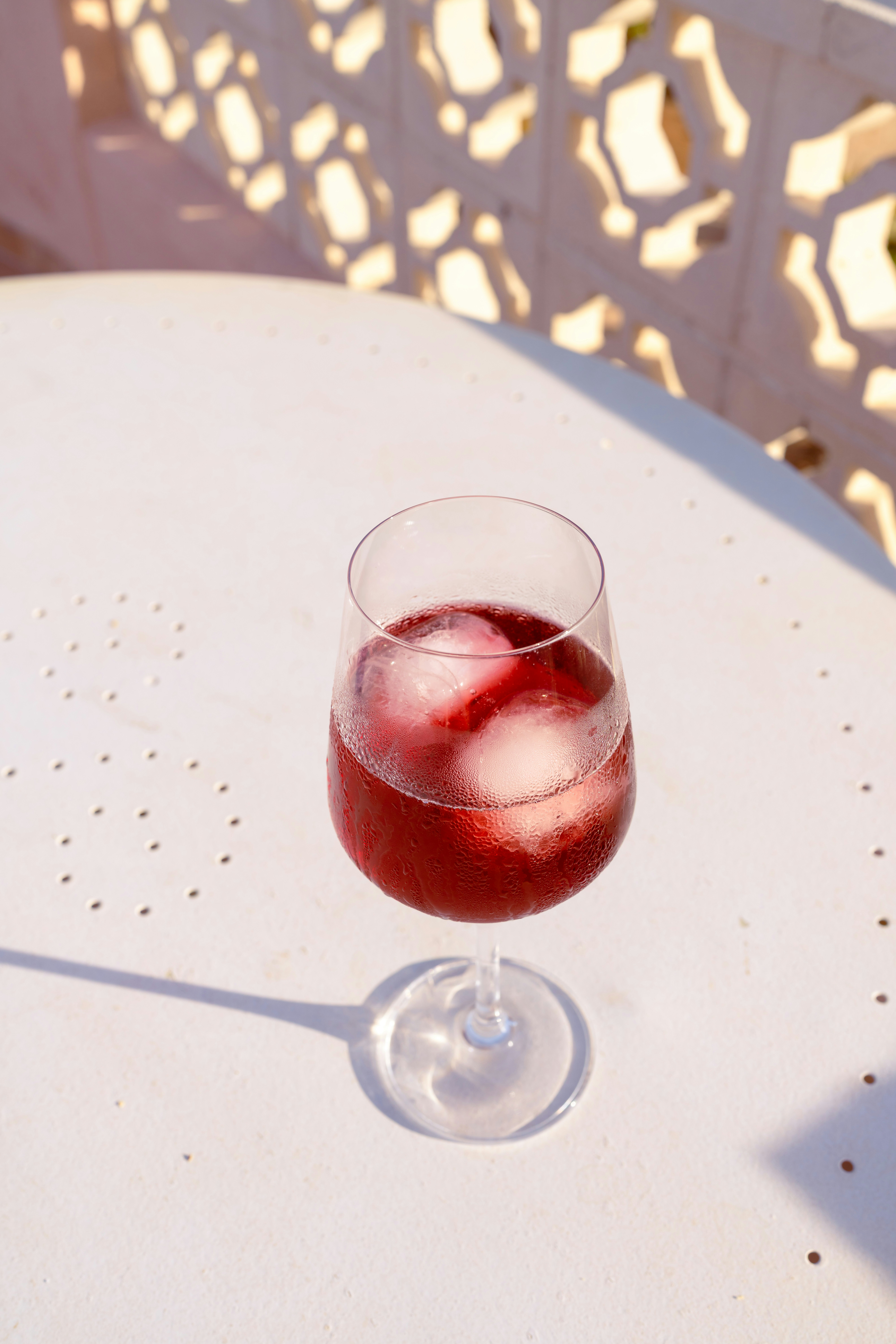 A glass of red iced beverage on a white perforated table in sunlight