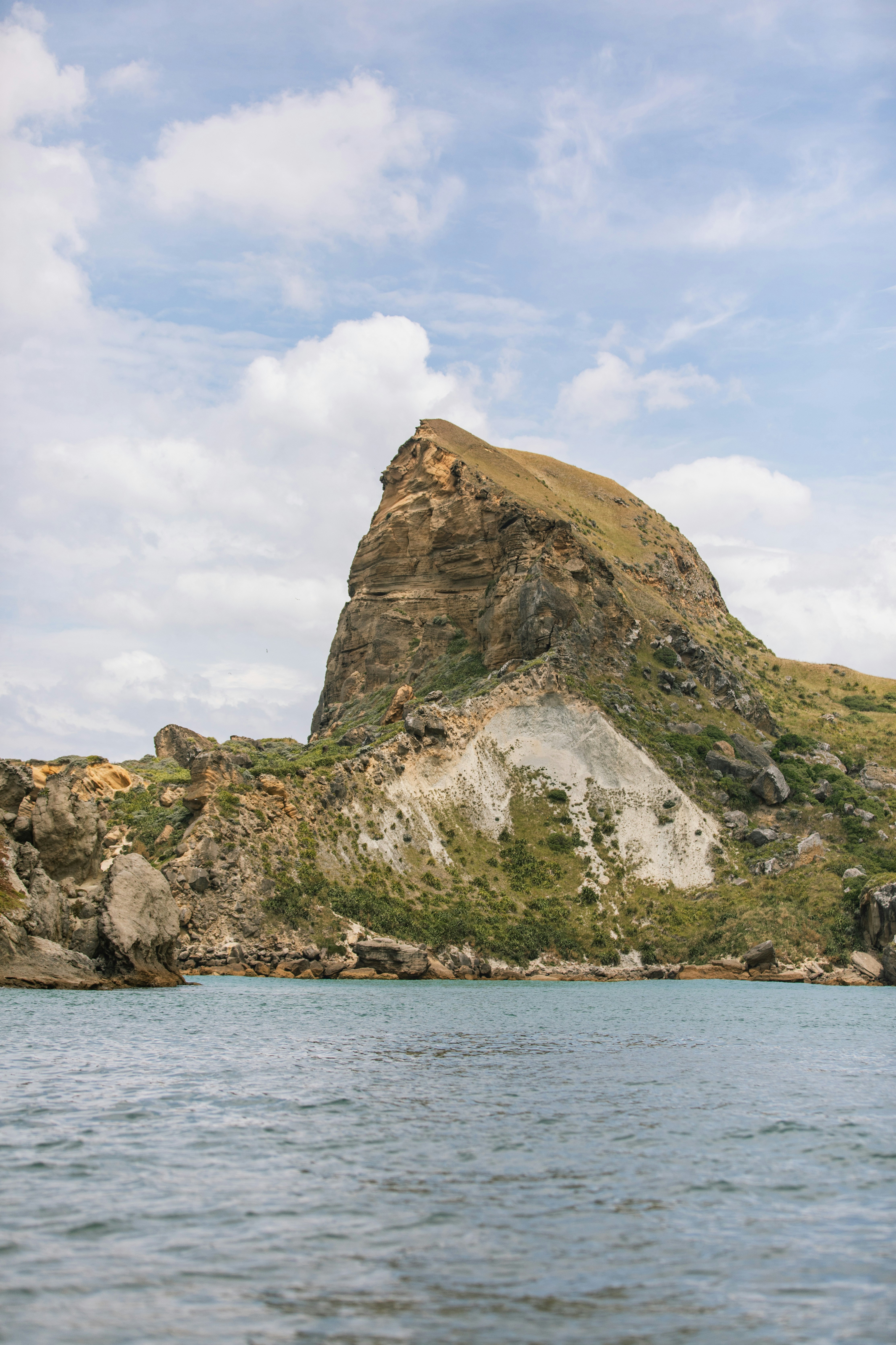 A steep rocky headland rising above calm blue water under a cloudy sky