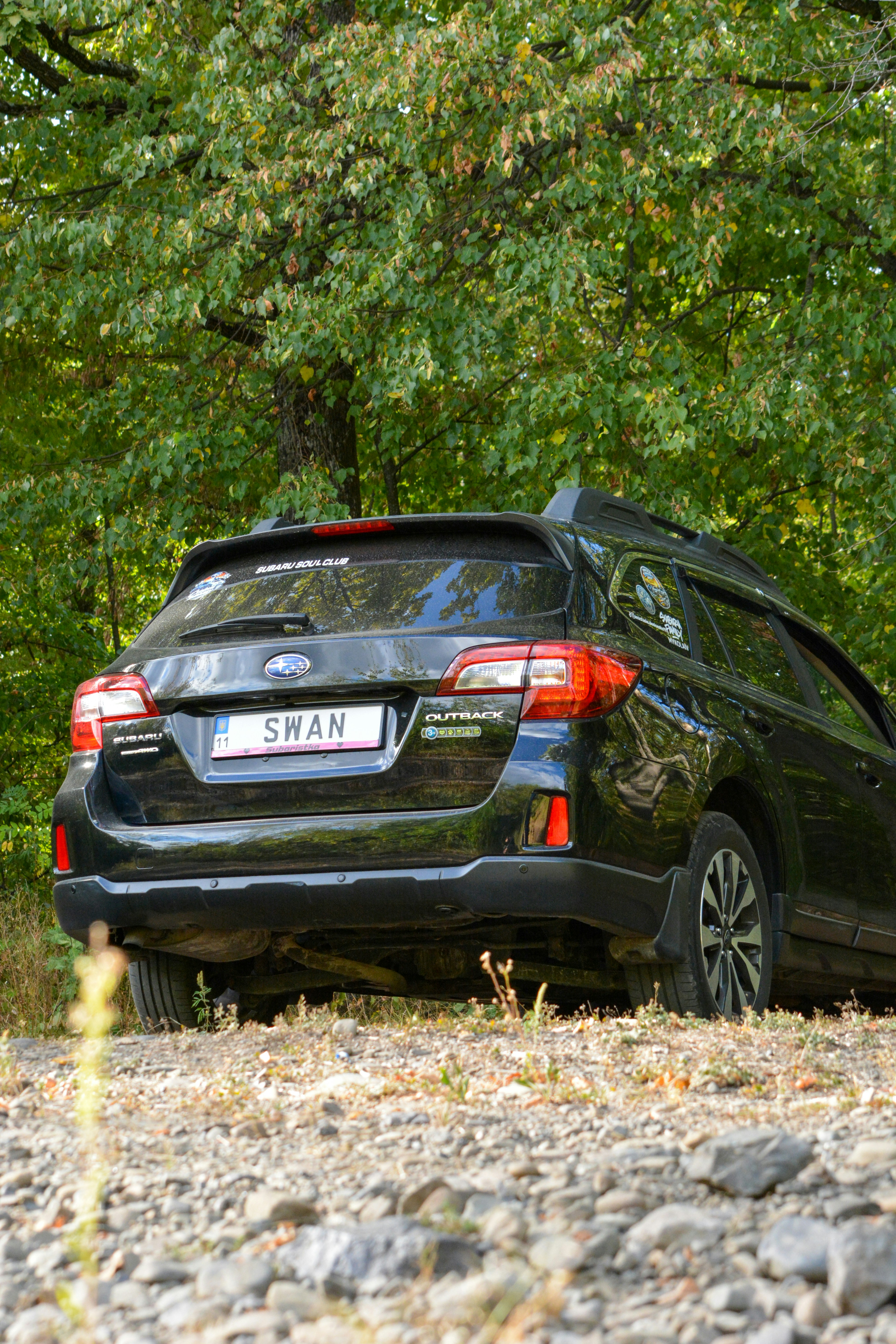 Black Subaru Outback parked on a gravel path before green trees
