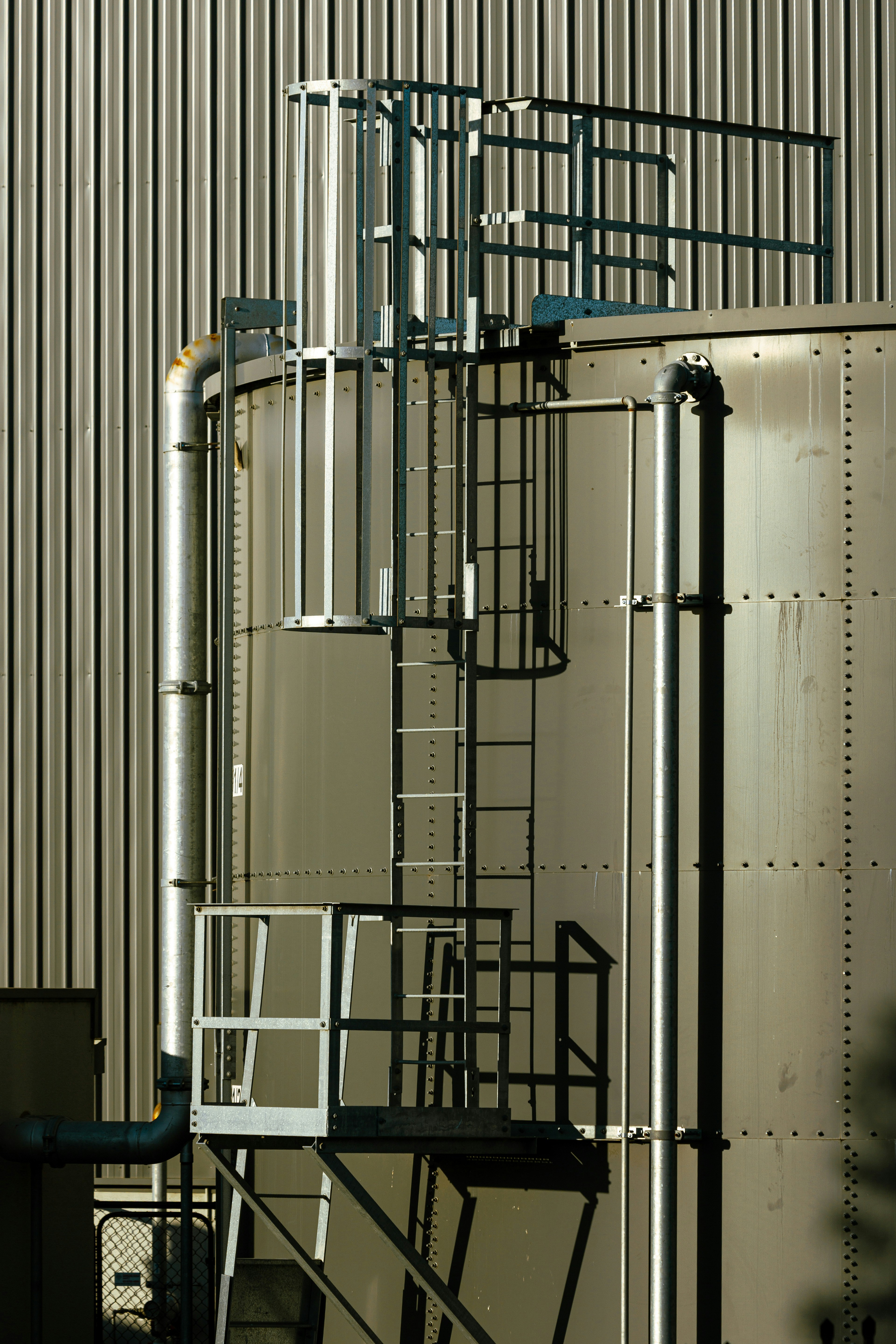 A metal storage tank with a caged ladder and platform against a corrugated wall