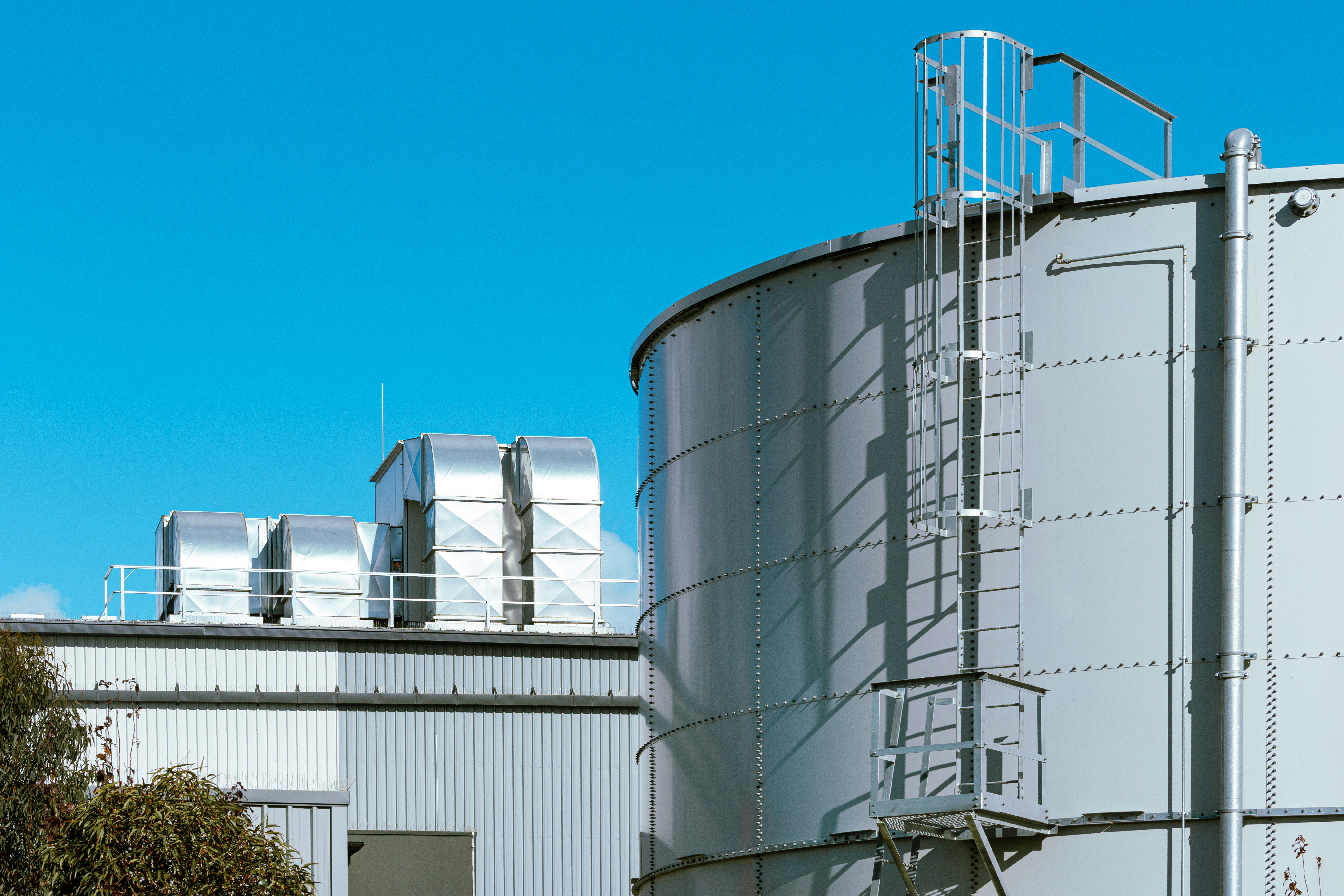 A large grey industrial storage tank with a metal ladder against a blue sky