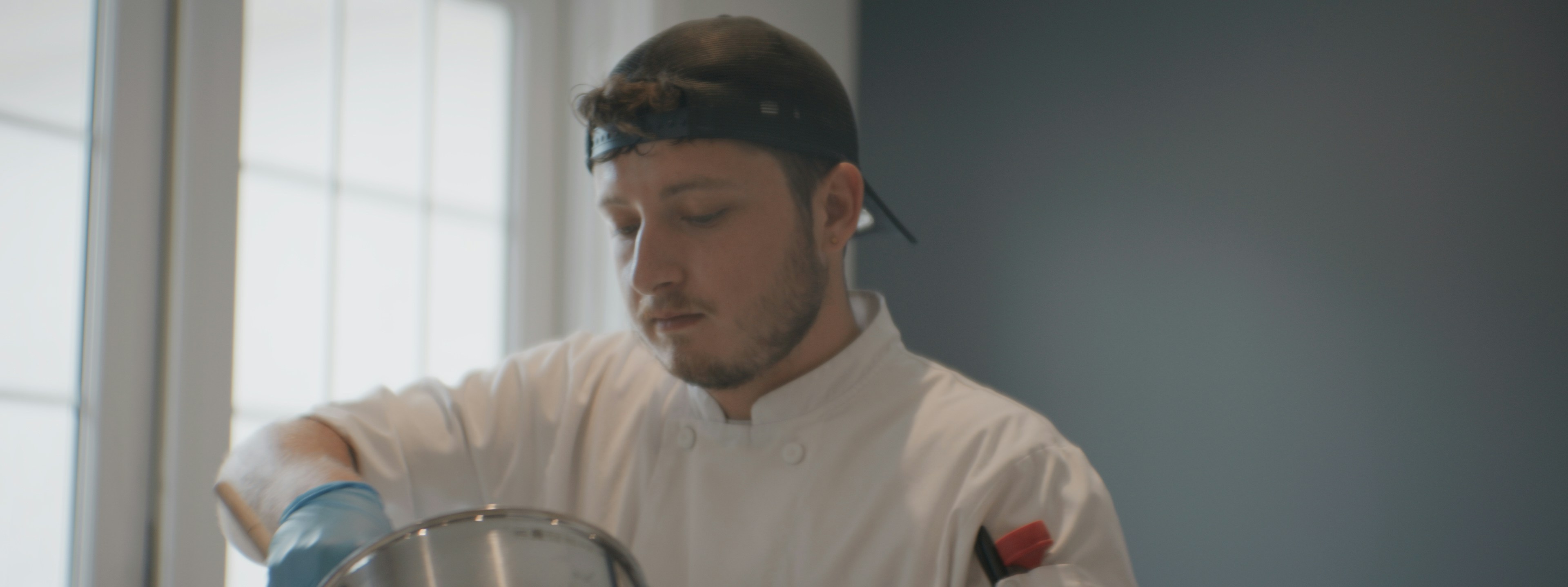 A person in a white chef coat and black cap stirring a metal pot