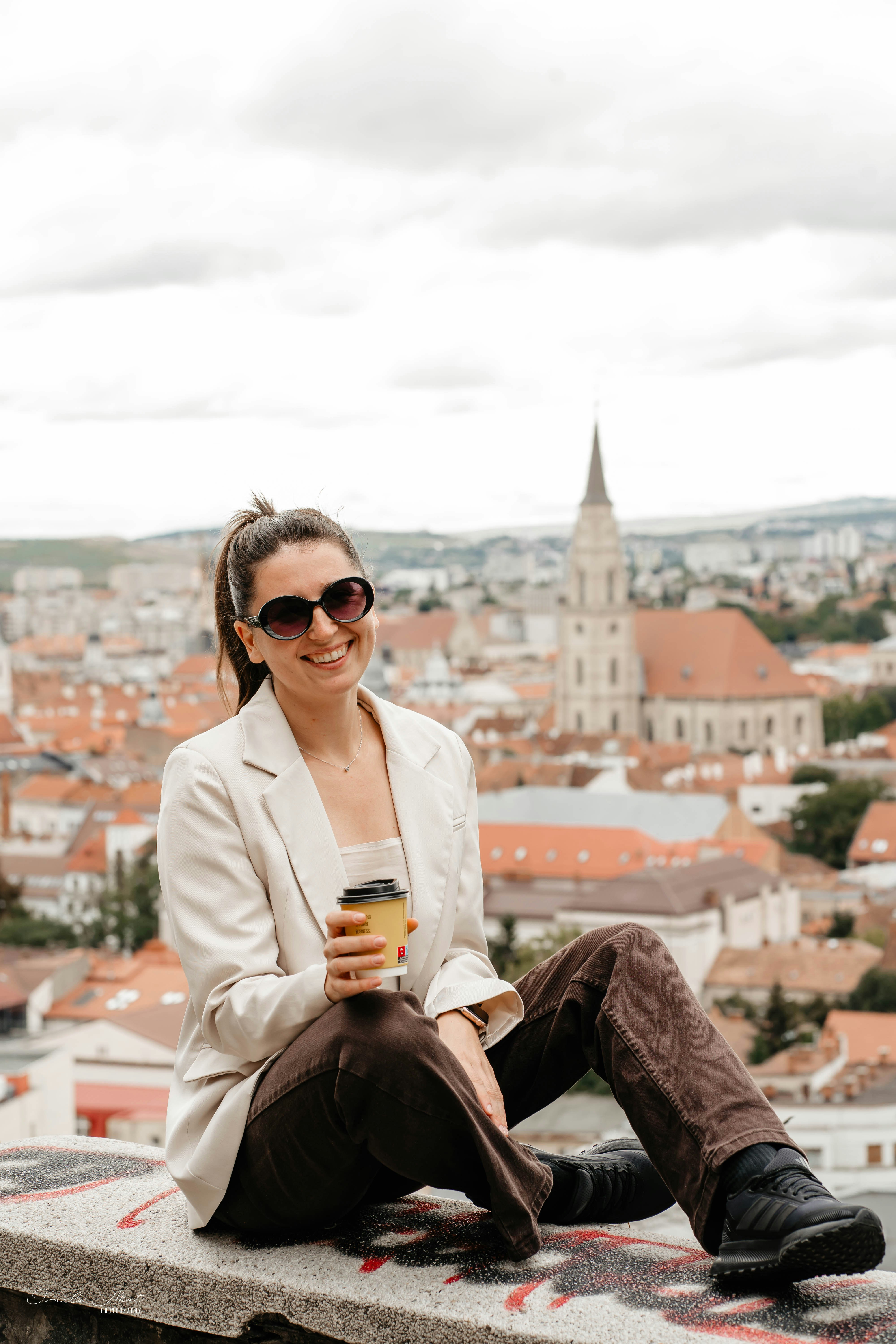 A woman in a beige blazer holding a coffee cup overlooking Cluj-Napoca