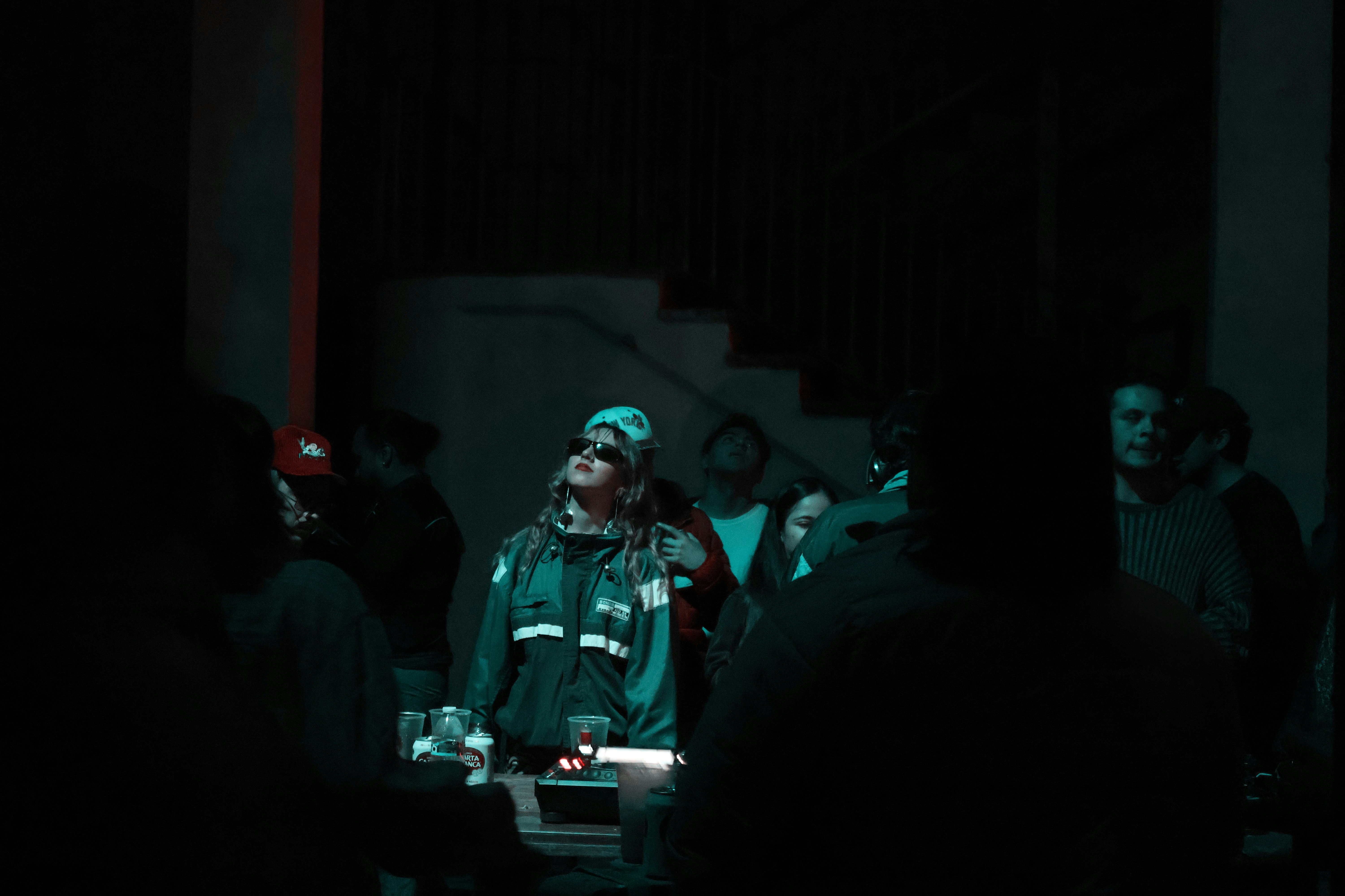 Dans une salle sombre, une femme en lunettes de soleil et veste verte regarde vers le haut, évoquant l’ambiance d’un club.