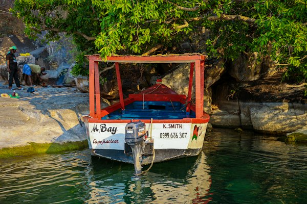 A boat at a rocky landing — A small motorboat moored under a leafy tree on Lake Malawi