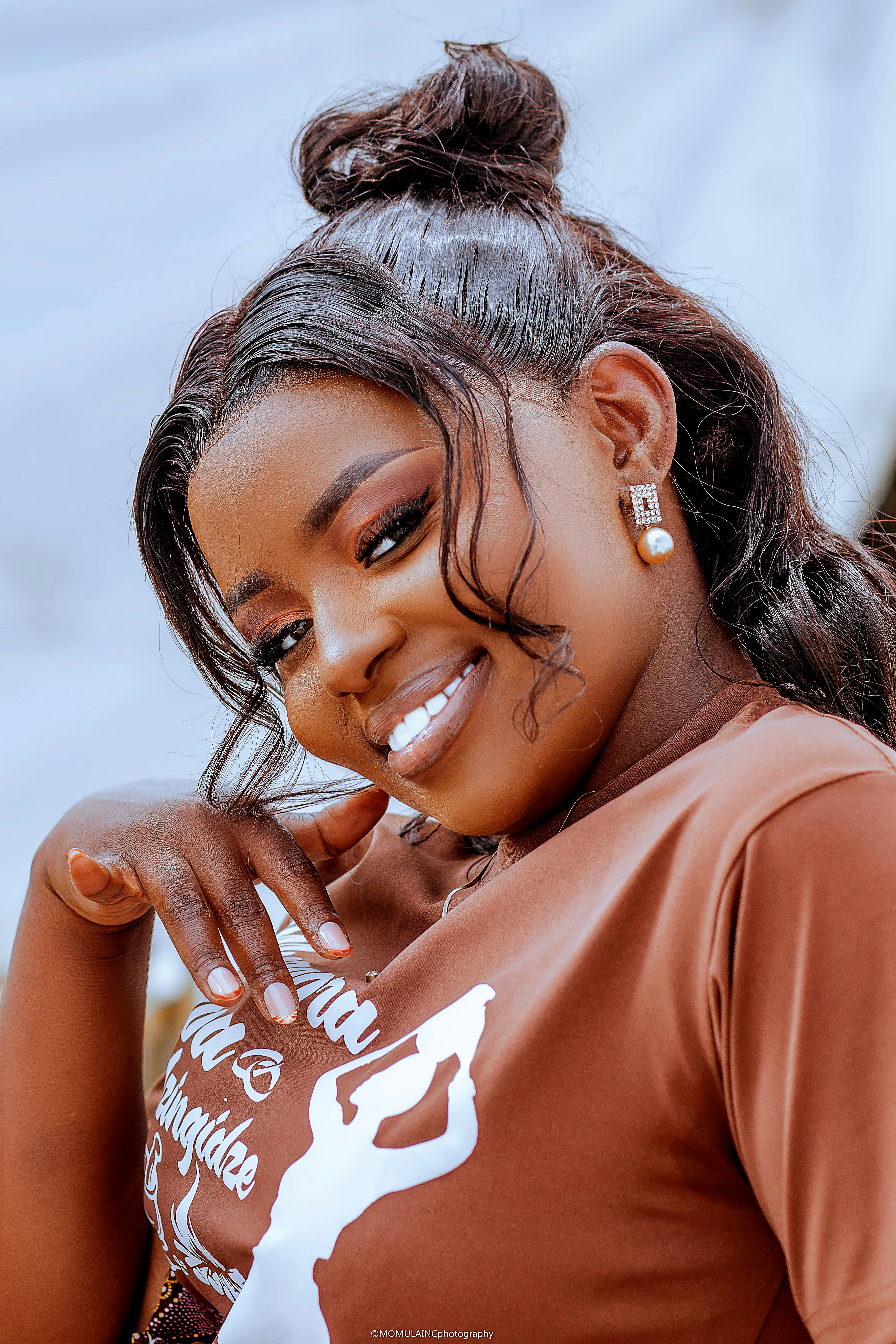 A smiling woman with hair in a bun wearing a brown t-shirt and pearl earrings