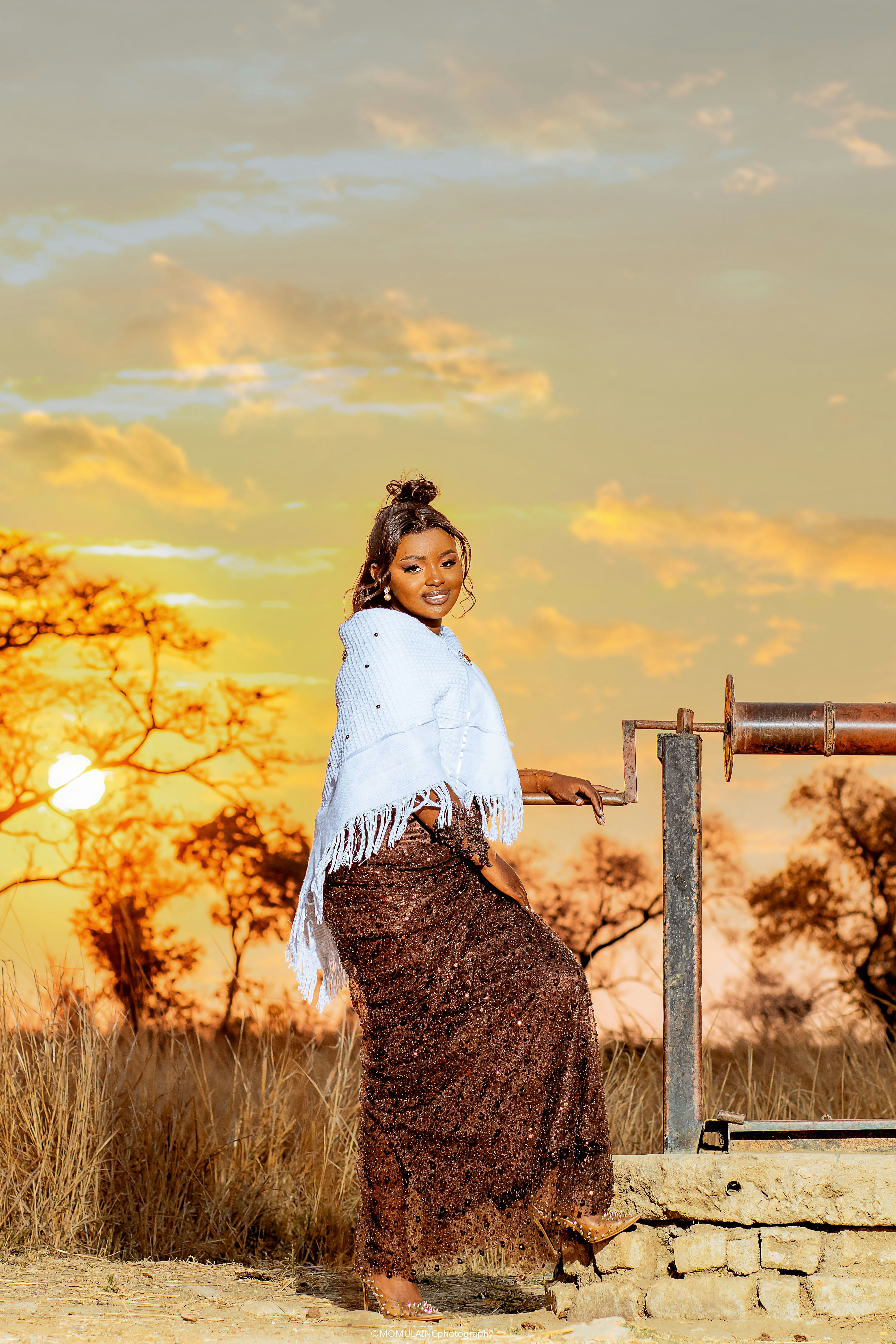 A woman in a sequined gown and white shawl beside a well at golden hour