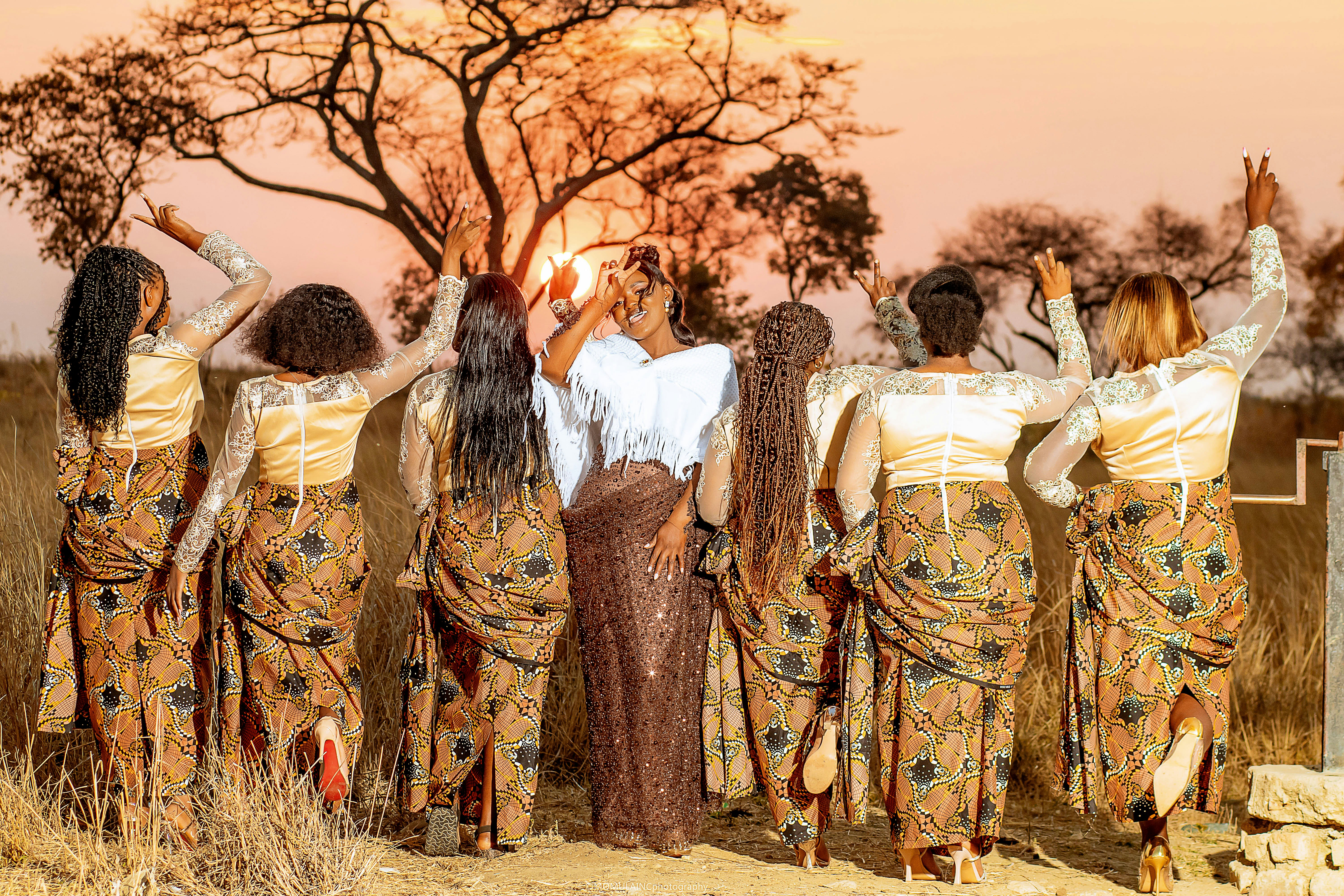 A group of women in patterned dresses posing under a warm orange sky