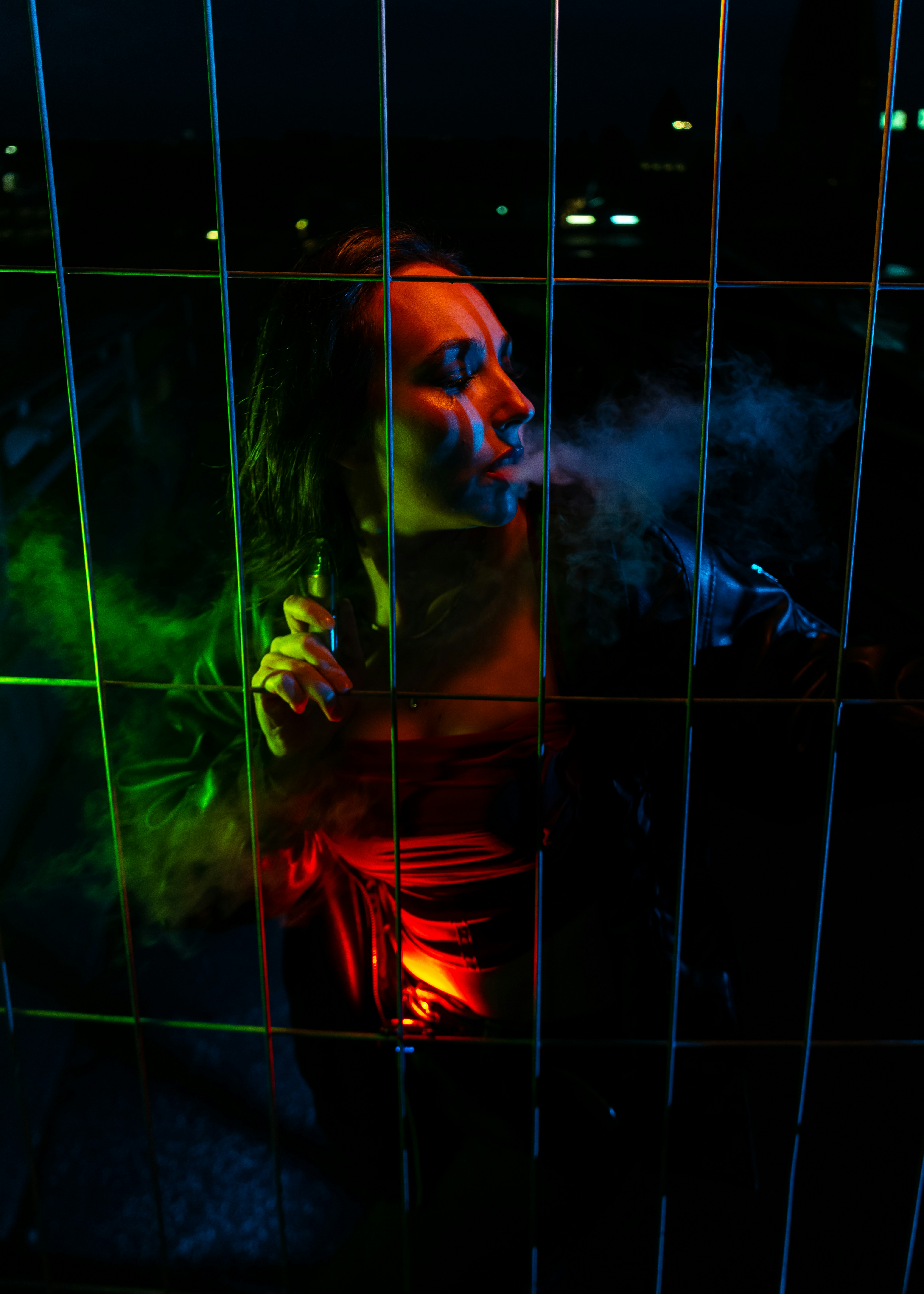 A woman exhaling vapor behind a metal fence under red and blue neon lights