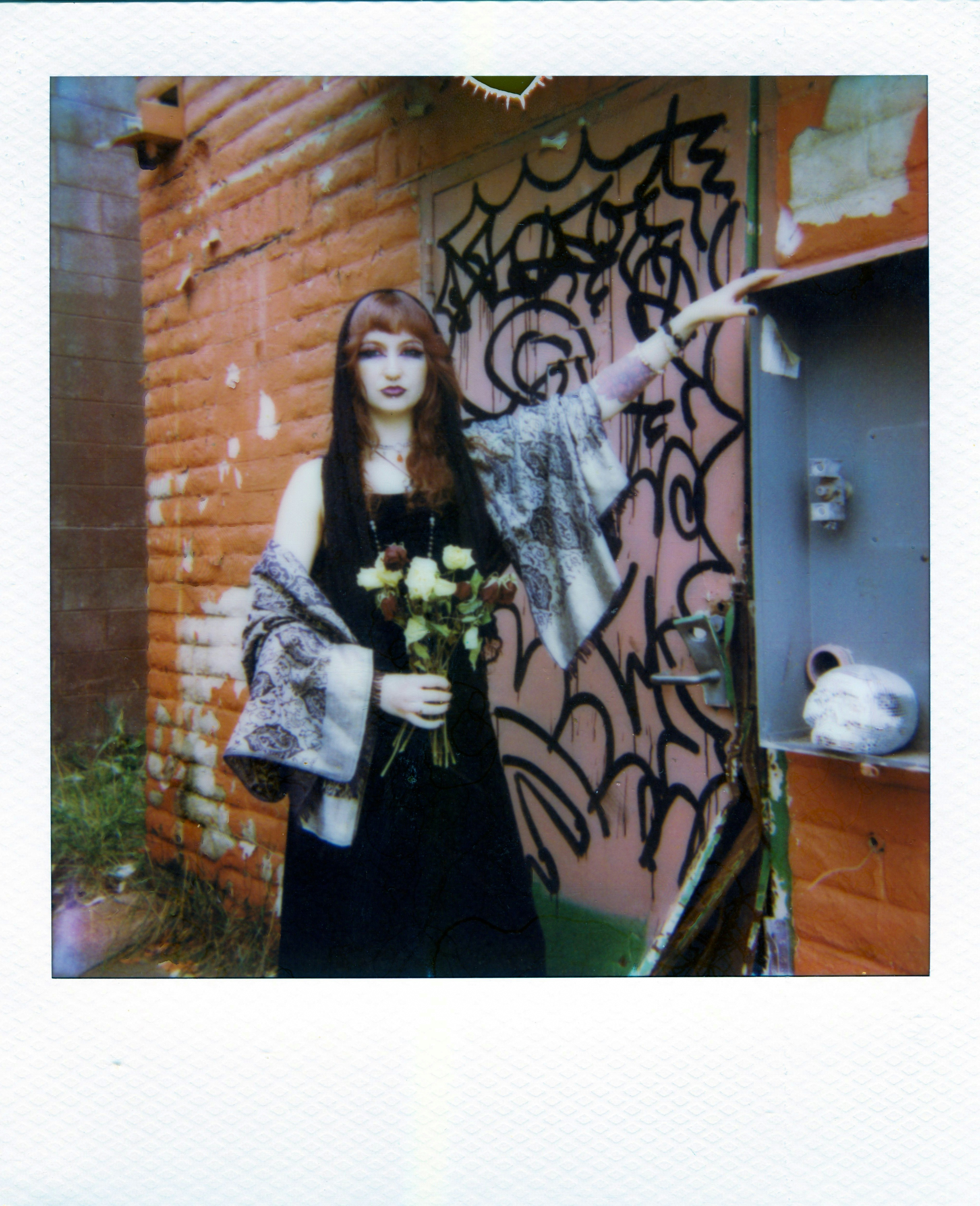 A woman in a black dress holding white roses against a graffiti covered wall