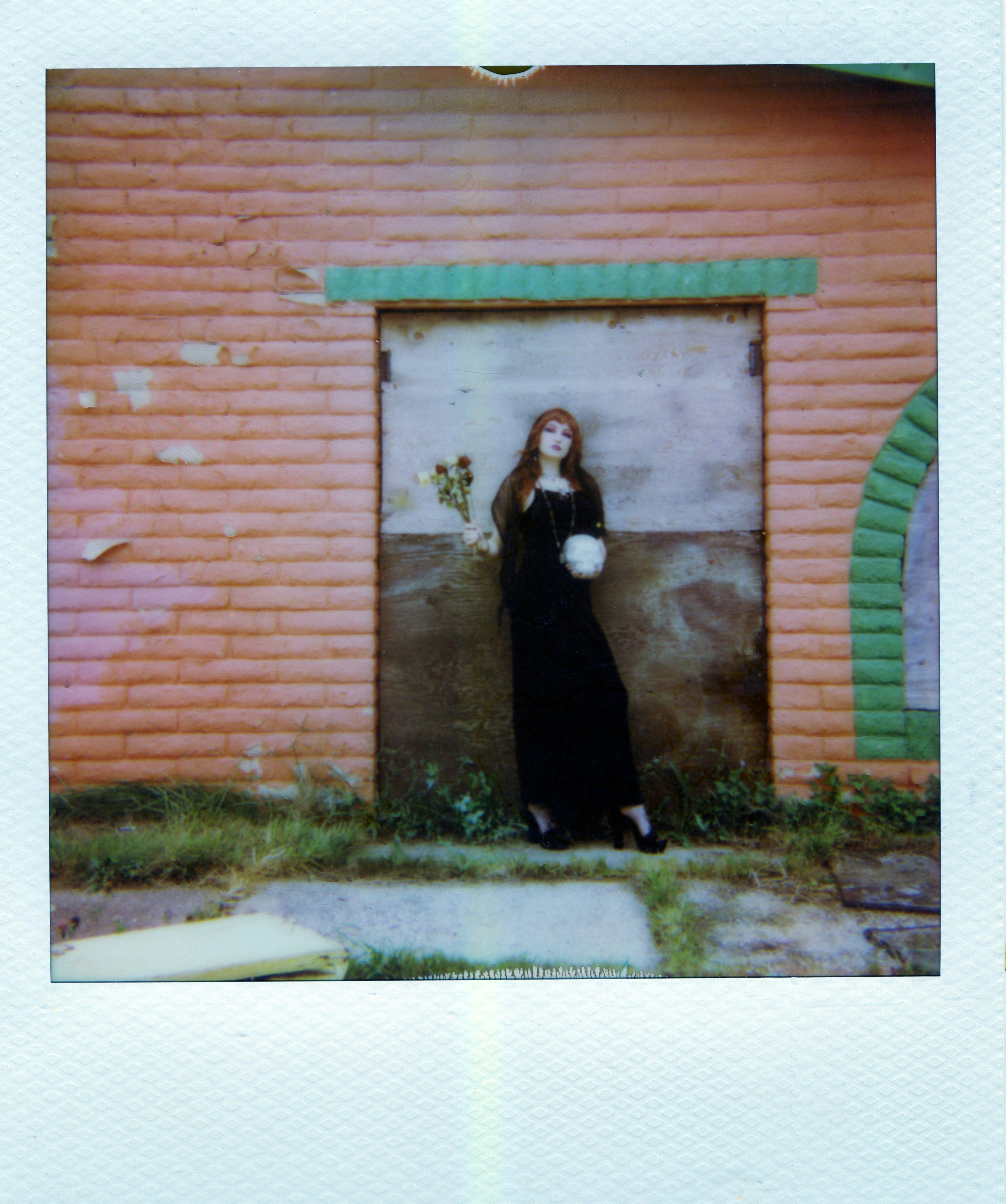 A person in a black dress holding flowers and a skull against a brick wall