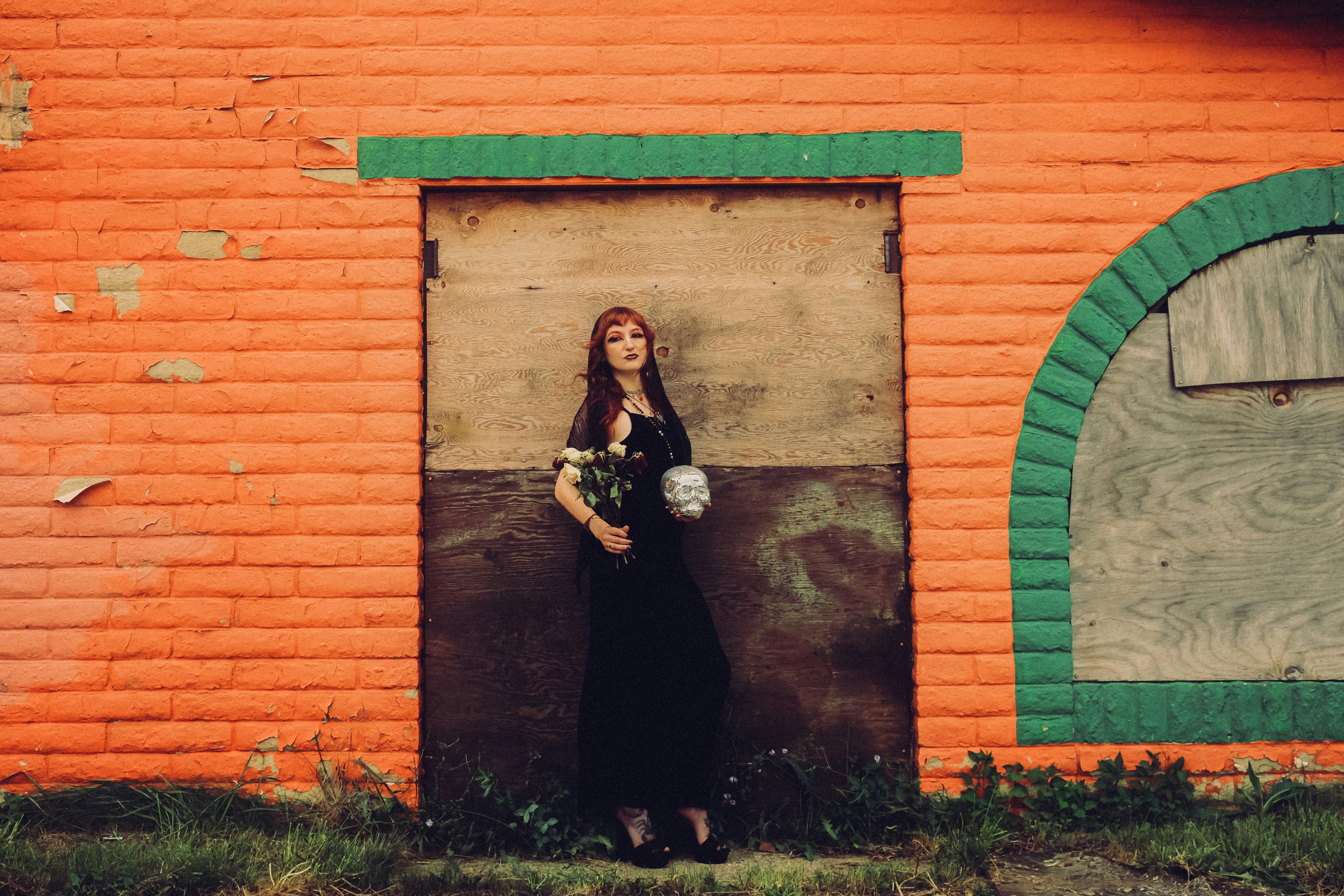 A woman in a black dress holding a bouquet and skull against an orange wall