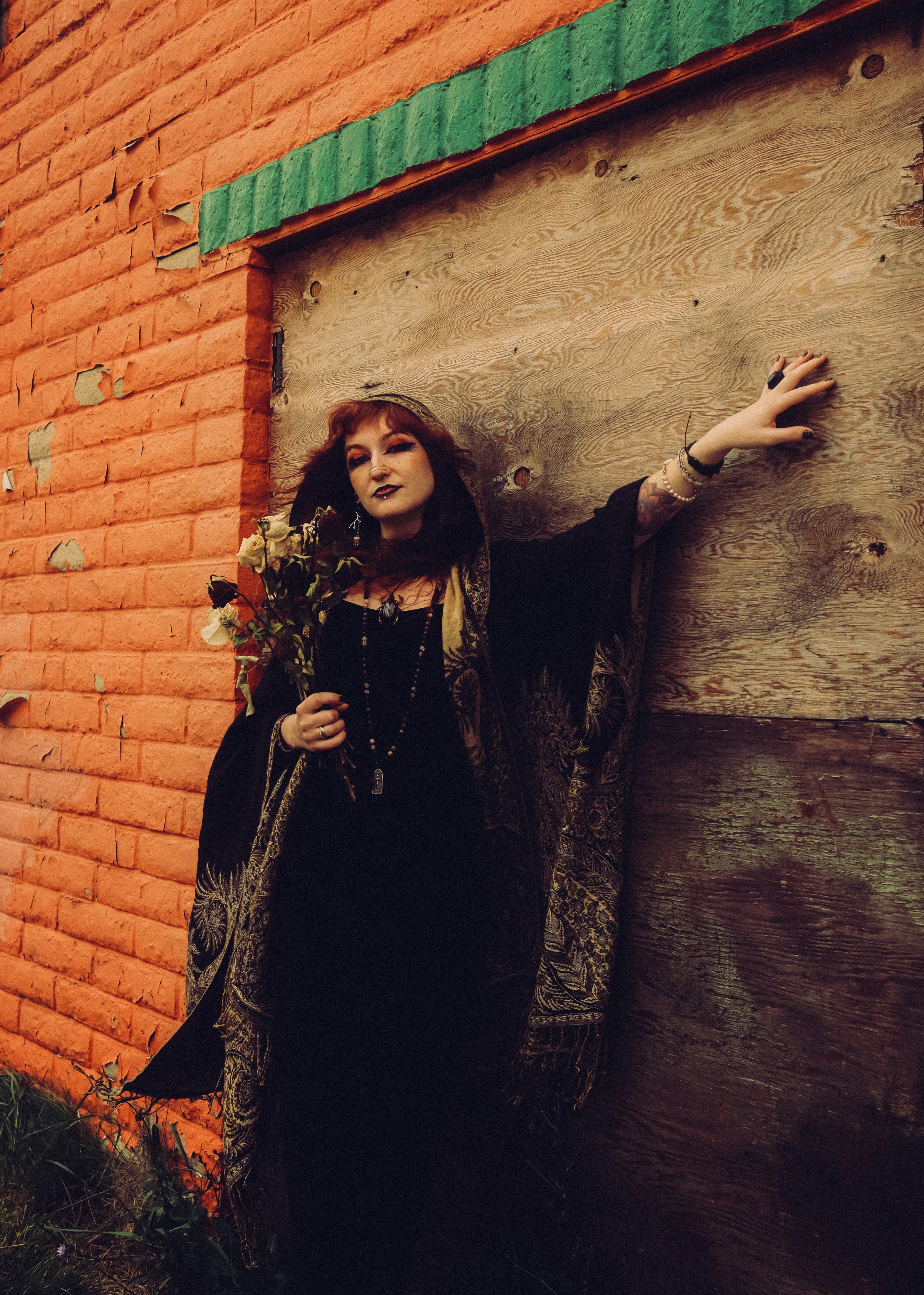 A person in a black hooded cloak holding dried flowers against an orange brick wall
