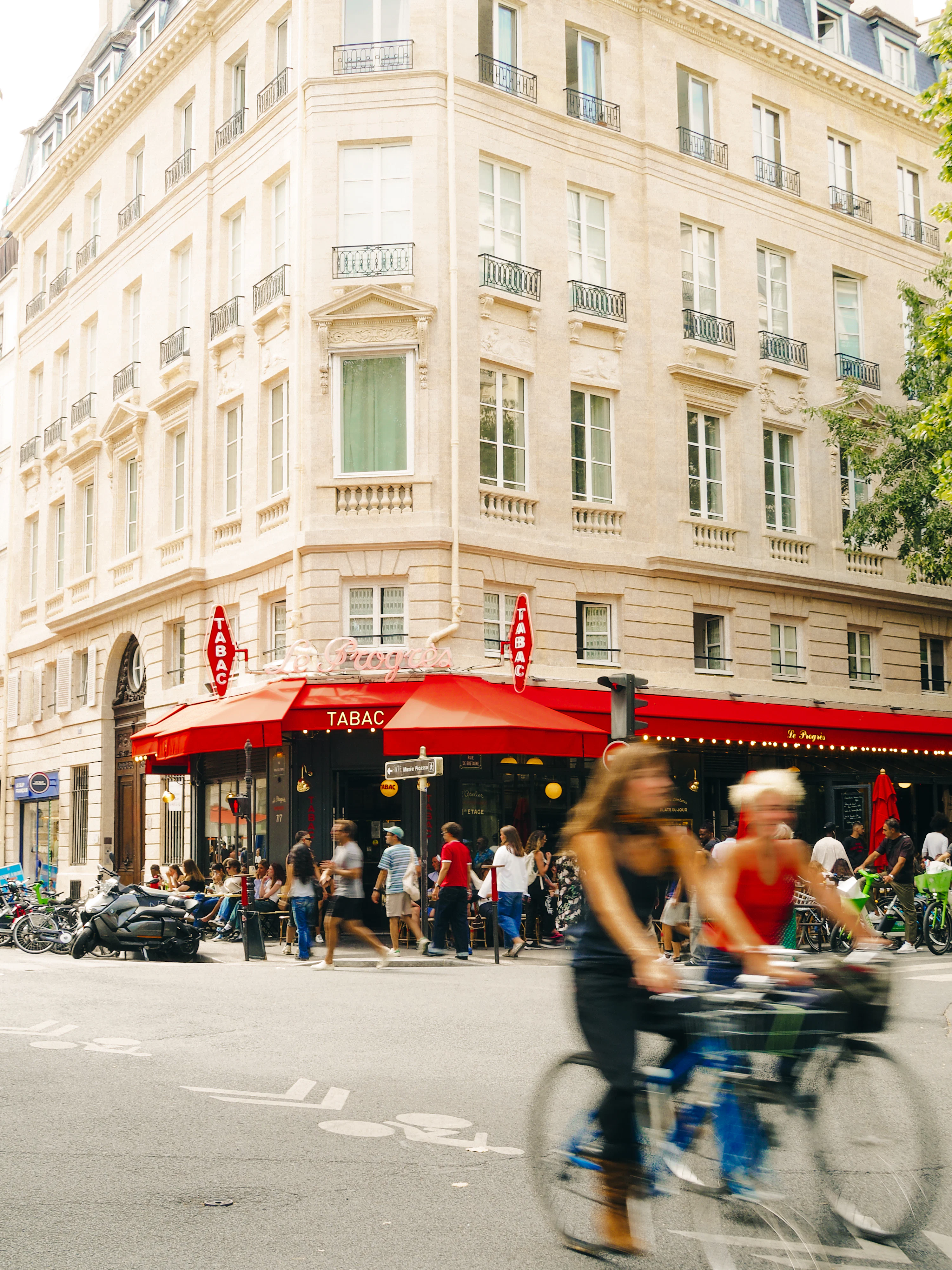 People cycling and walking past a corner cafe with red awnings in Paris