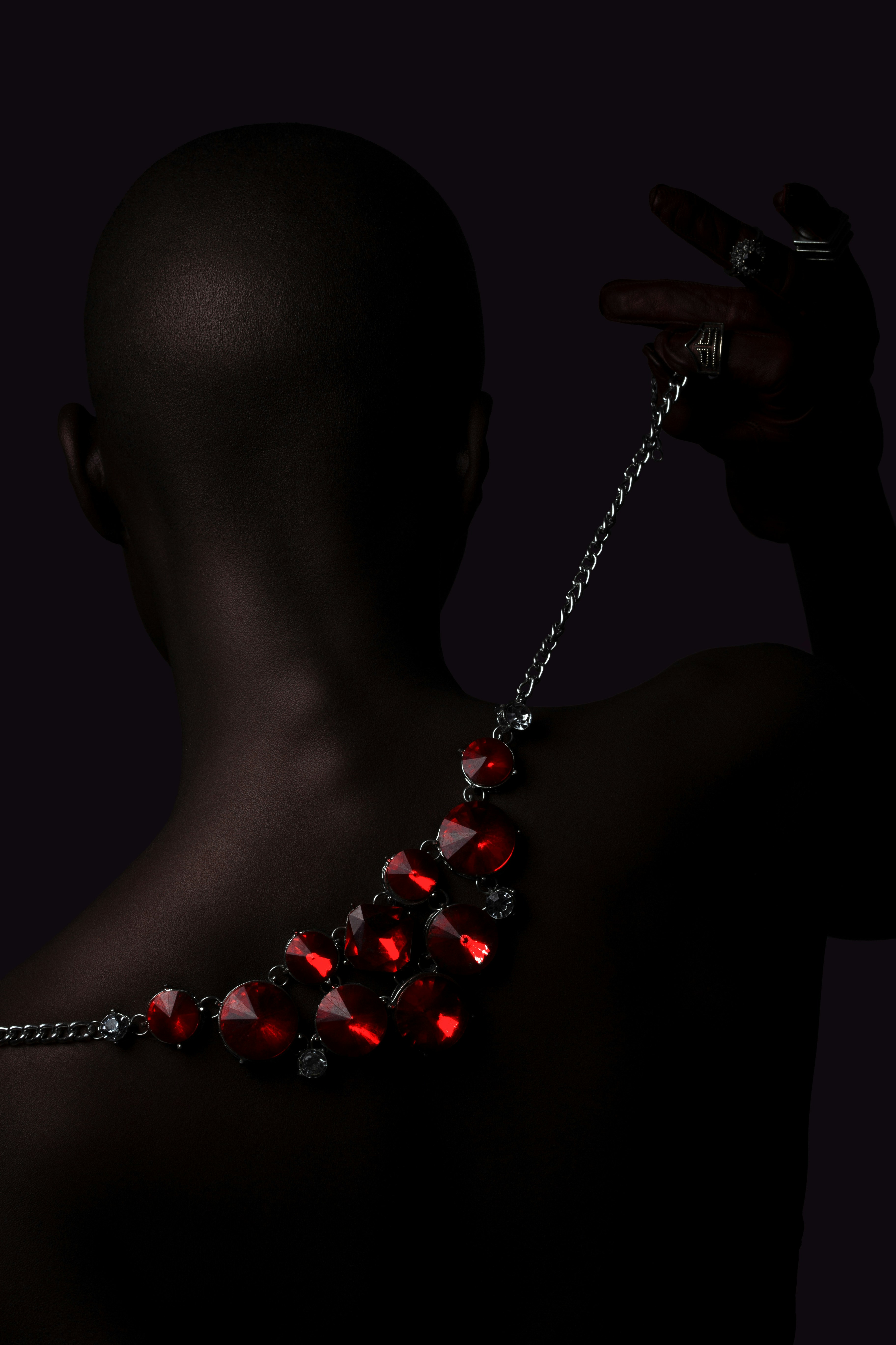 A person with a shaved head wearing a red crystal necklace against dark background