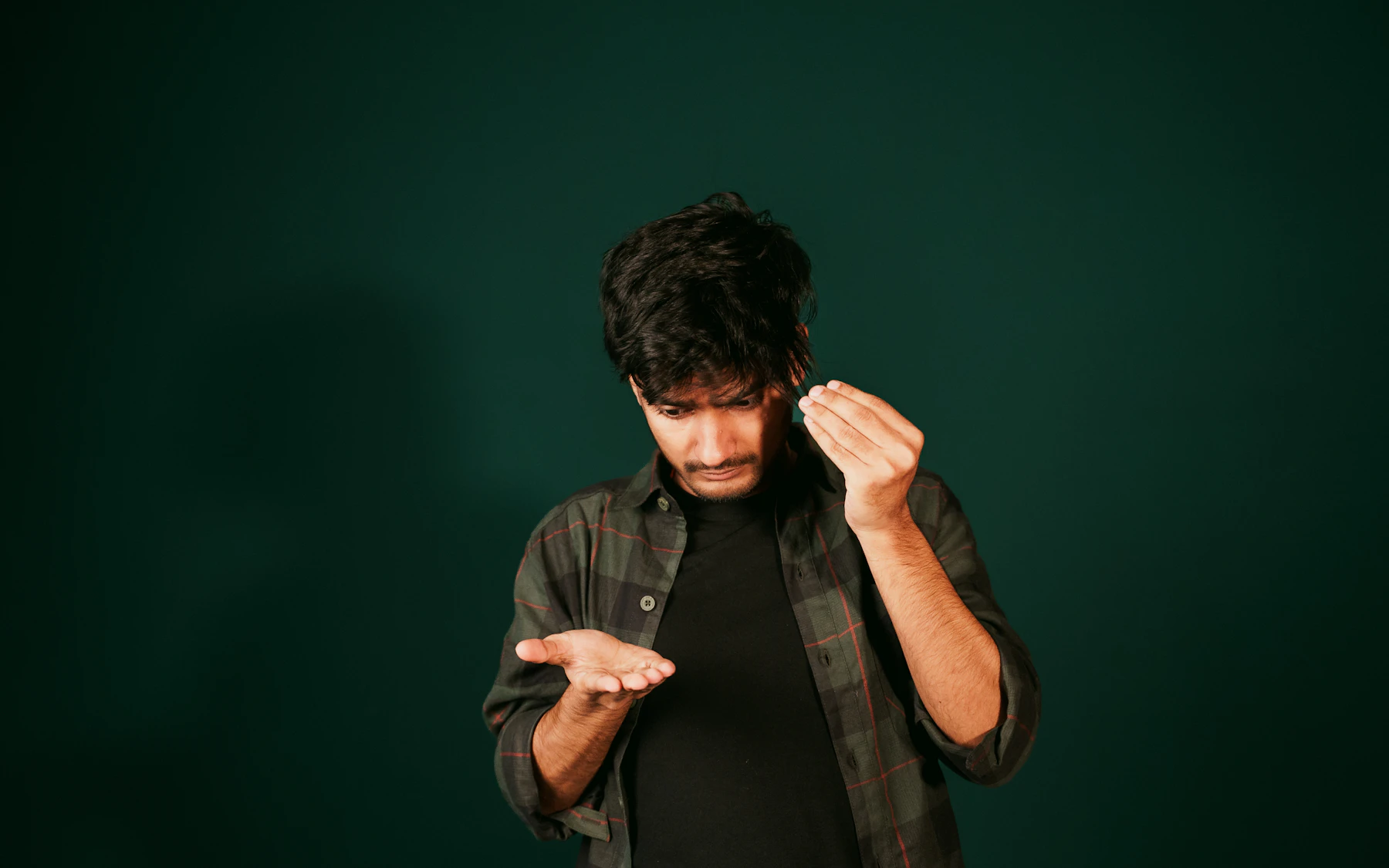 A man in a plaid shirt looking down at his open palm, his other hand raised beside his head, against a dark green wall