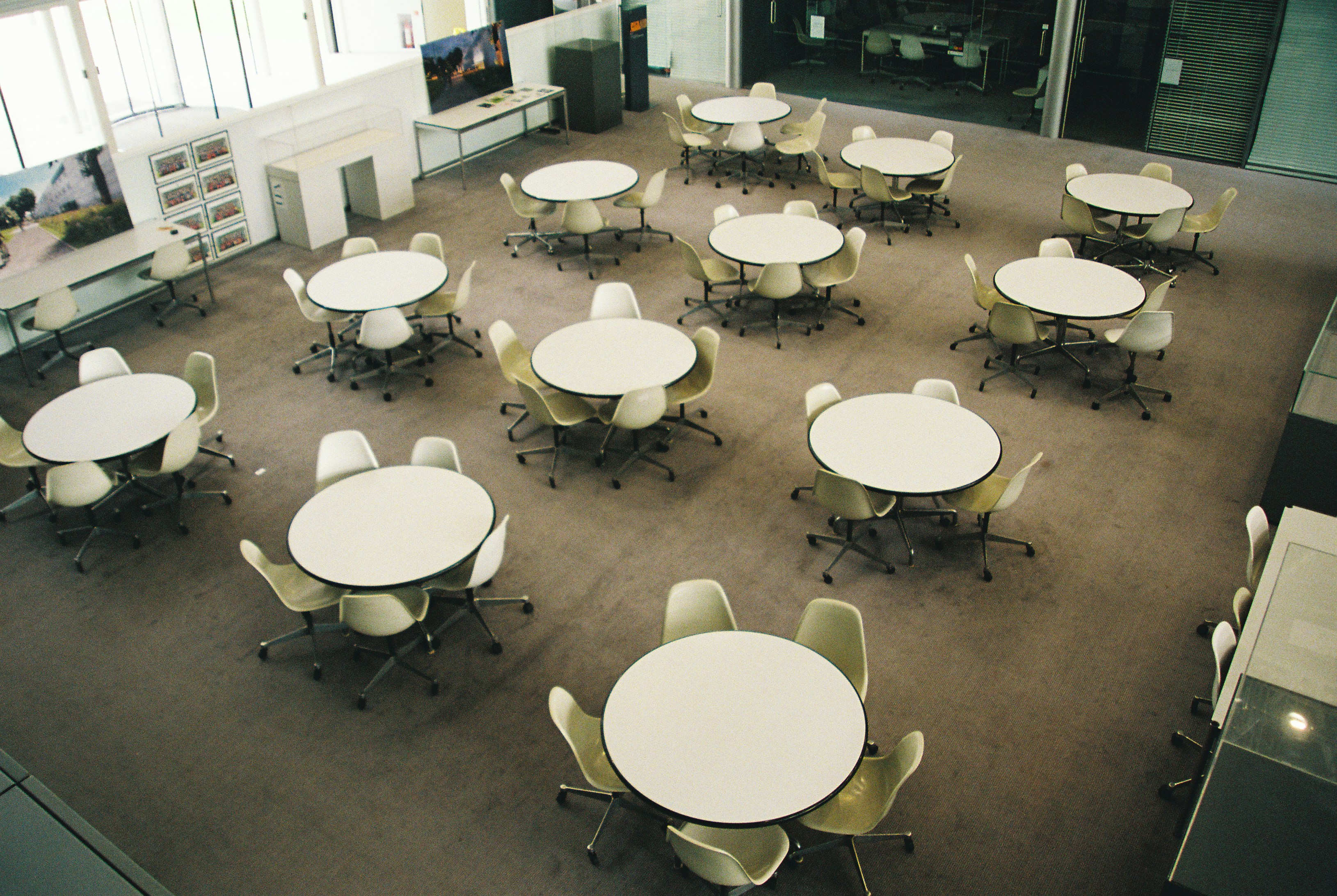 Rows of round tables and swivel chairs sit empty in a bright university common room, seen from above.