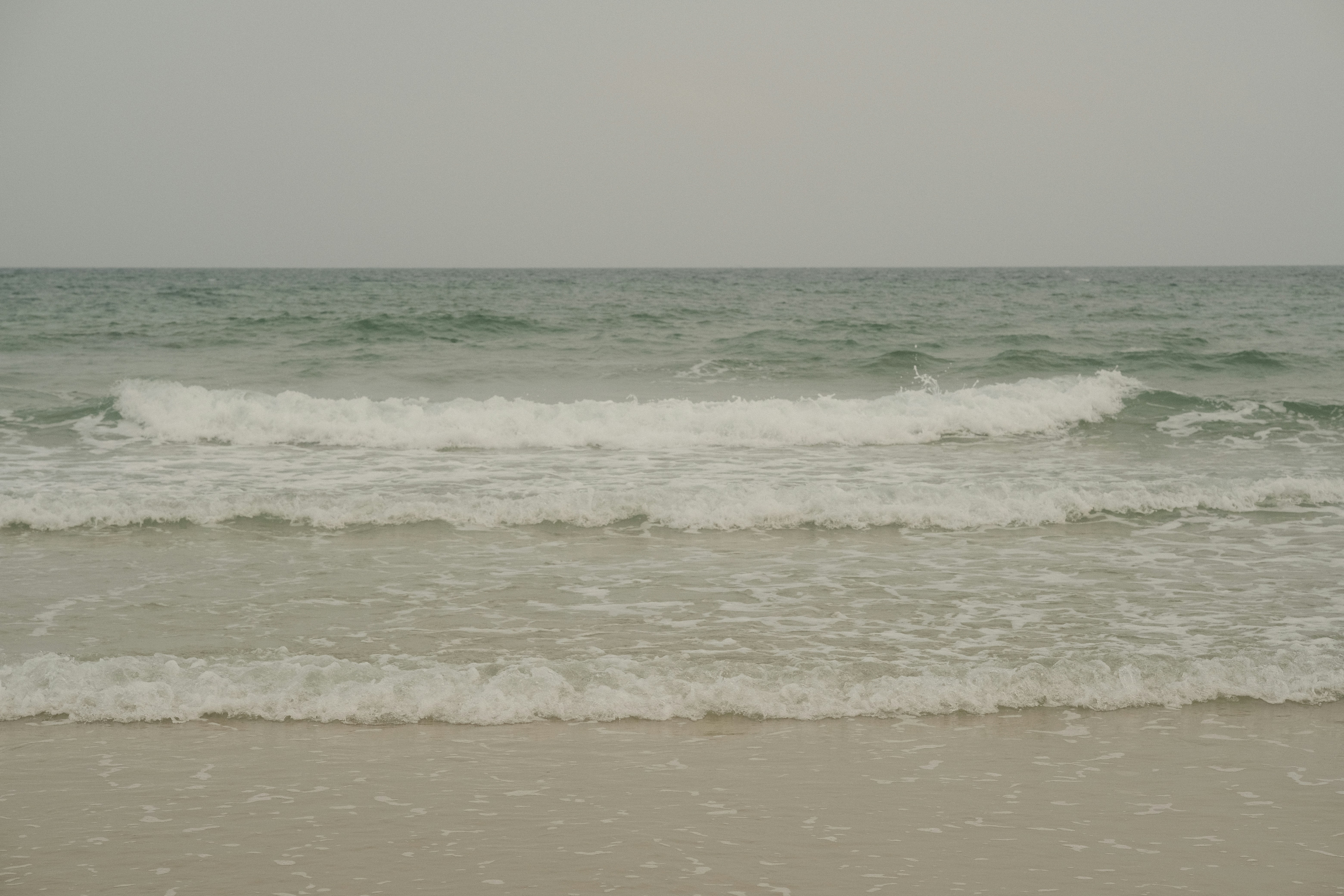 Three rows of gentle white waves rolling onto a sandy beach under overcast skies