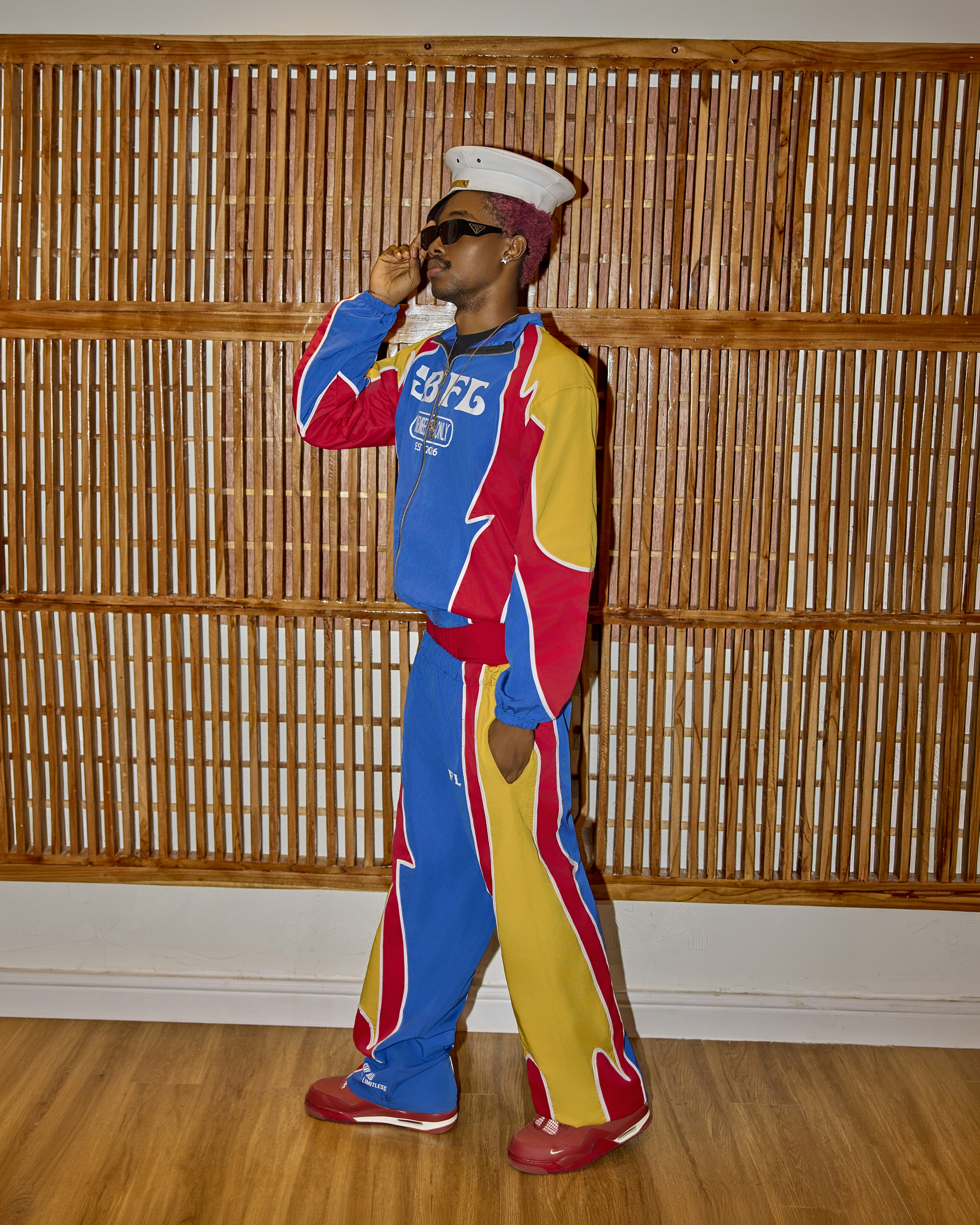 A person in a colorful tracksuit and sailor hat against a wooden slat wall