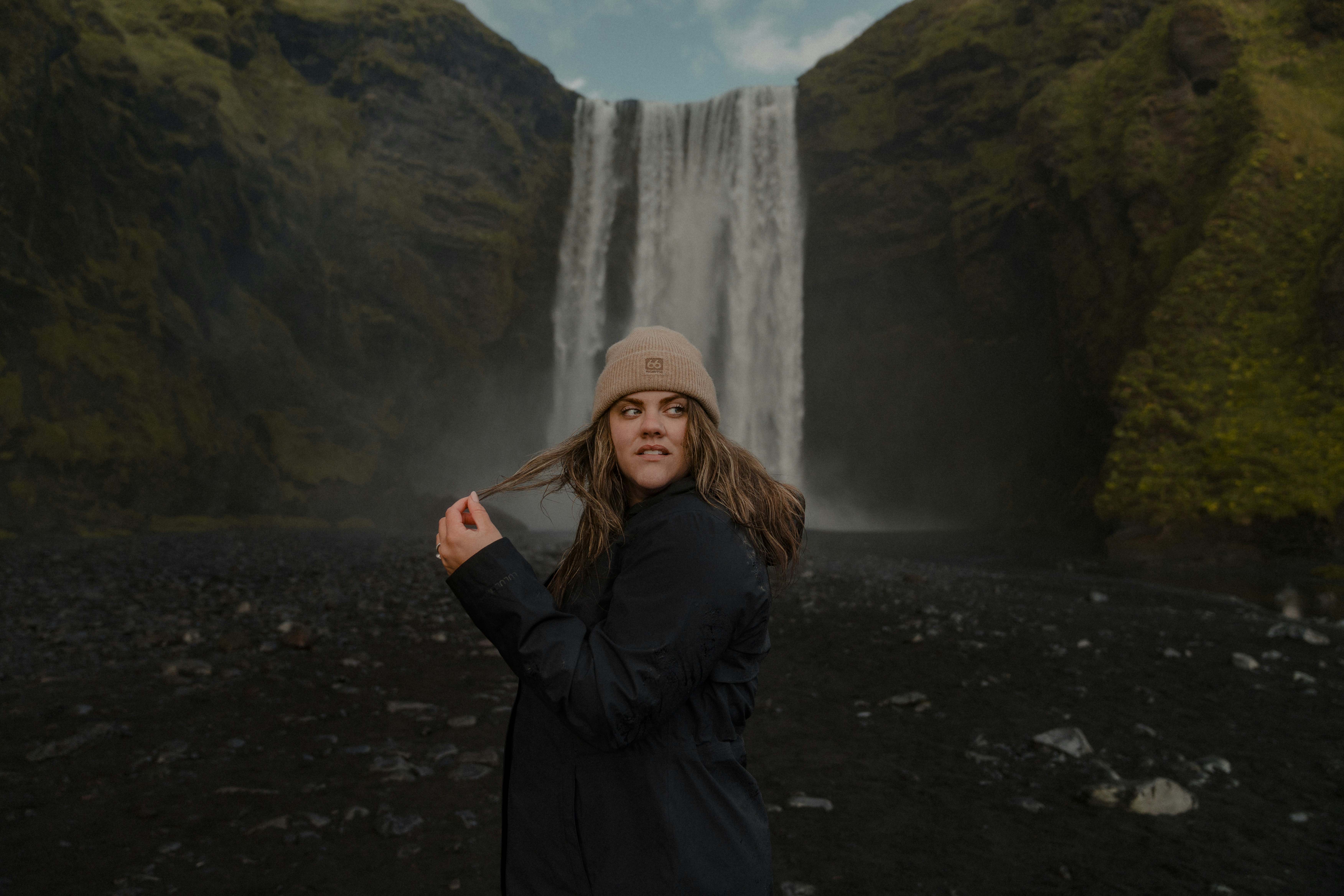 A woman in a black jacket and beanie standing before a large waterfall