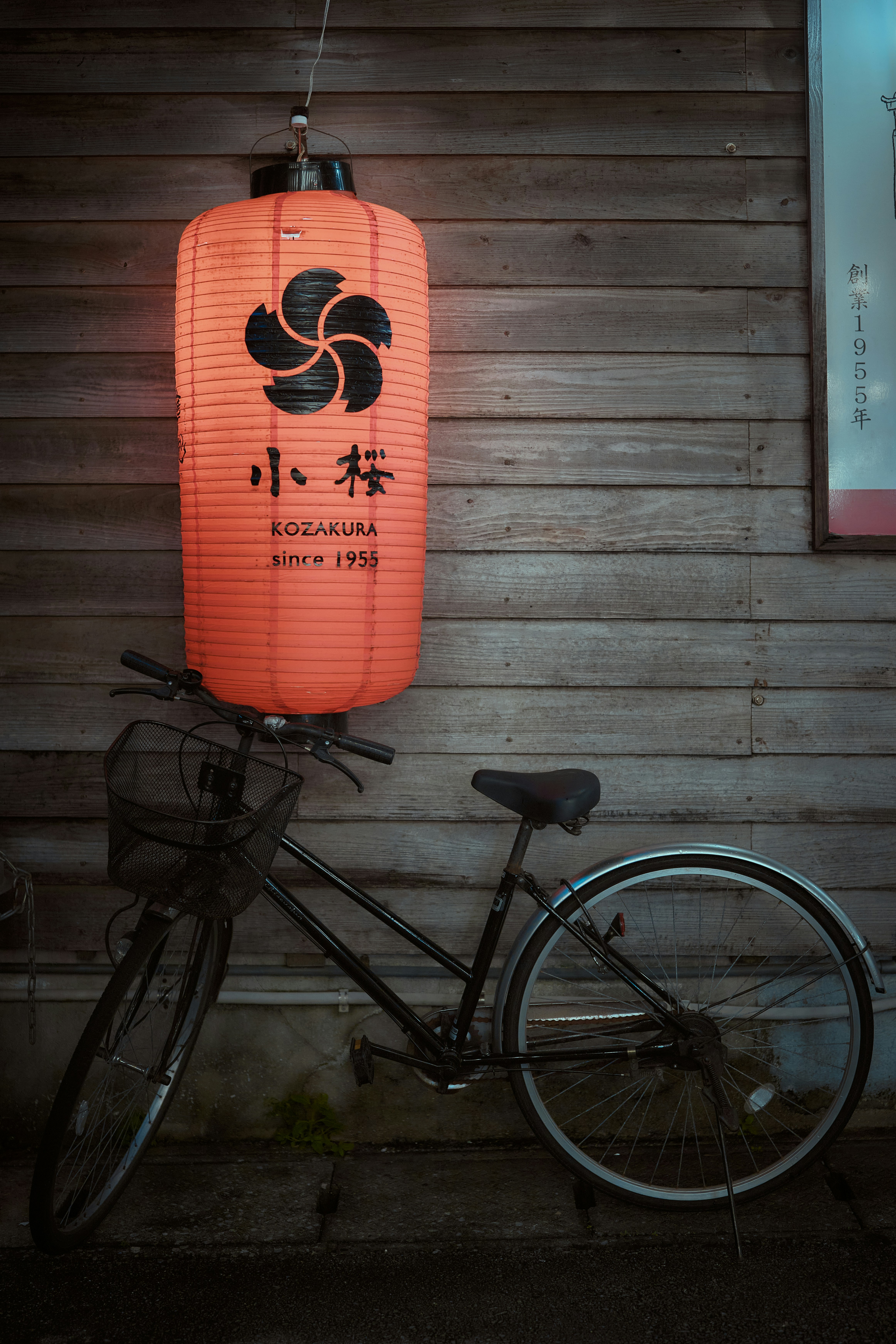 A black bicycle parked against a wooden wall beneath an orange paper lantern in Naha