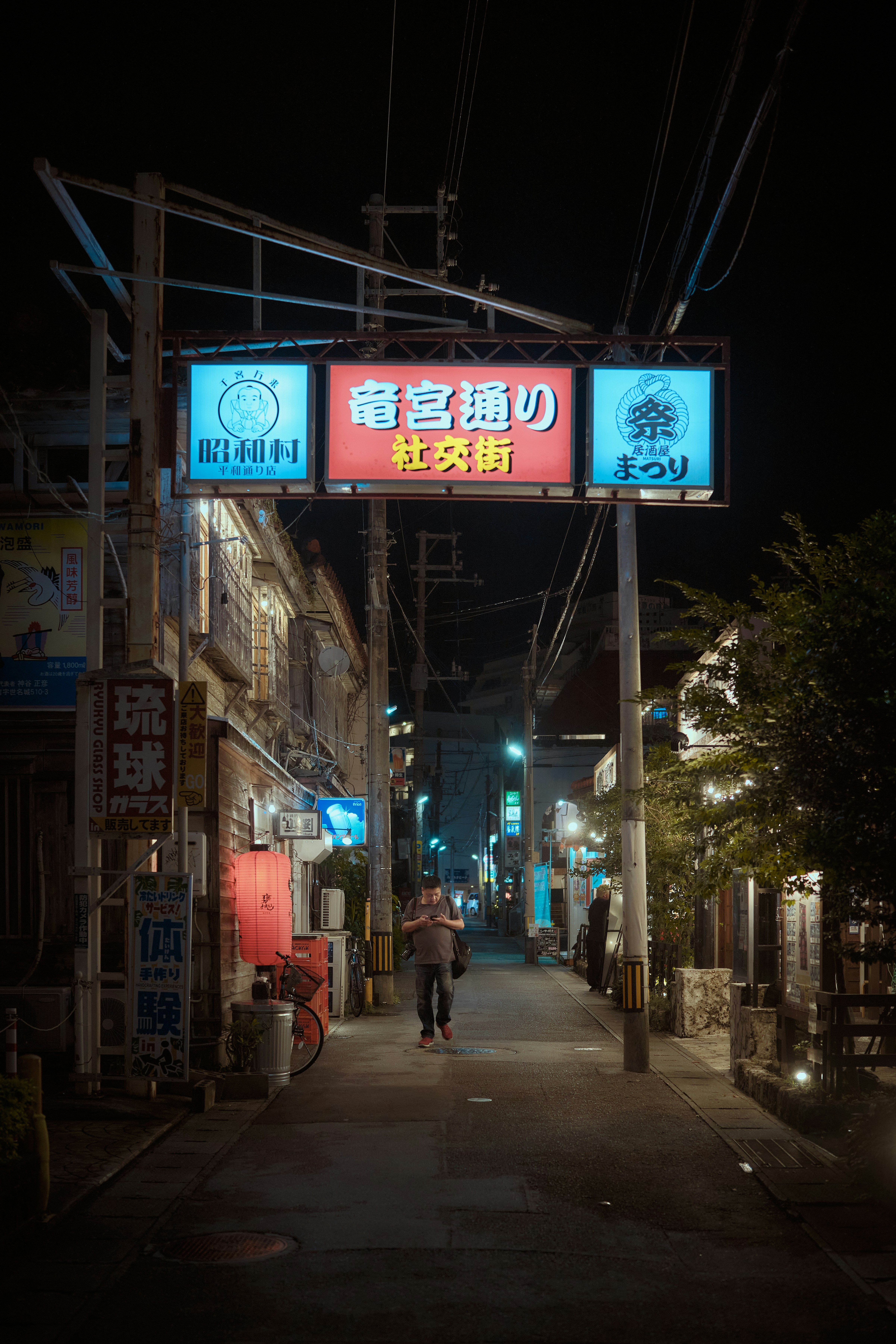 A person walking down a narrow street at night under illuminated signs in Naha