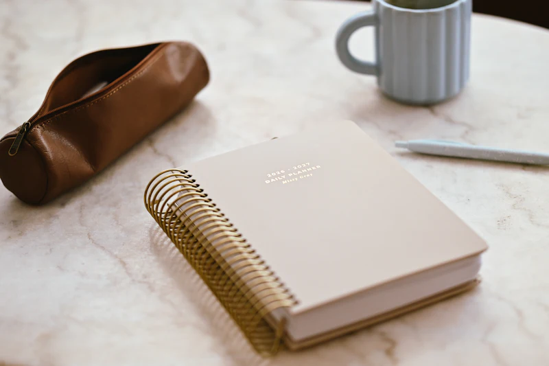 A beige planner, pencil case, and mug on a marble table