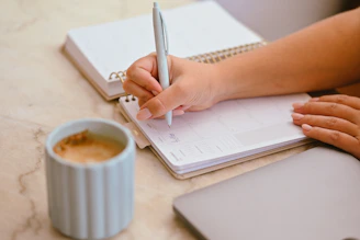 A person writing in a planner with a light blue pen near a coffee mug