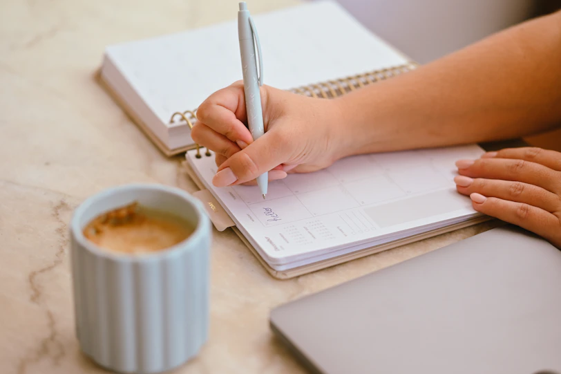 A person writing in a planner with a light blue pen near a coffee mug