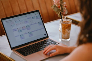 A person using a laptop with a calendar interface beside an iced coffee