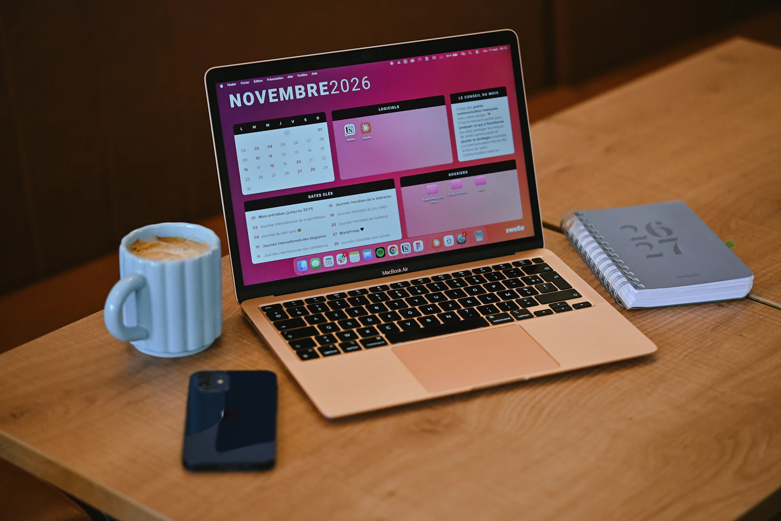 A laptop showing a monthly calendar on a wooden desk next to a coffee mug, a phone and a notebook