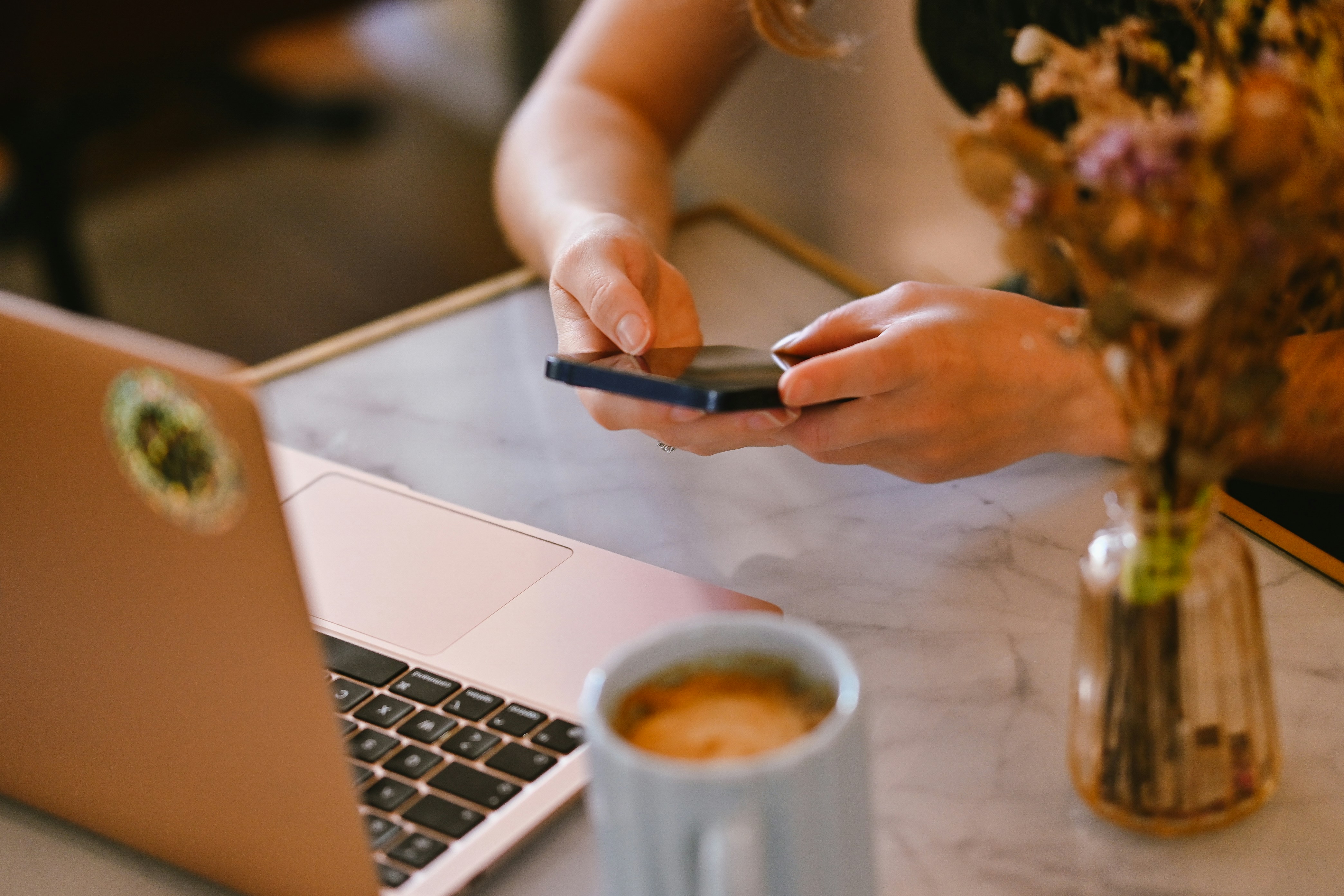 A person holding a smartphone at a marble table with a laptop and flowers