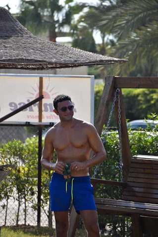 A shirtless man in blue swim trunks and sunglasses holding a snack bag