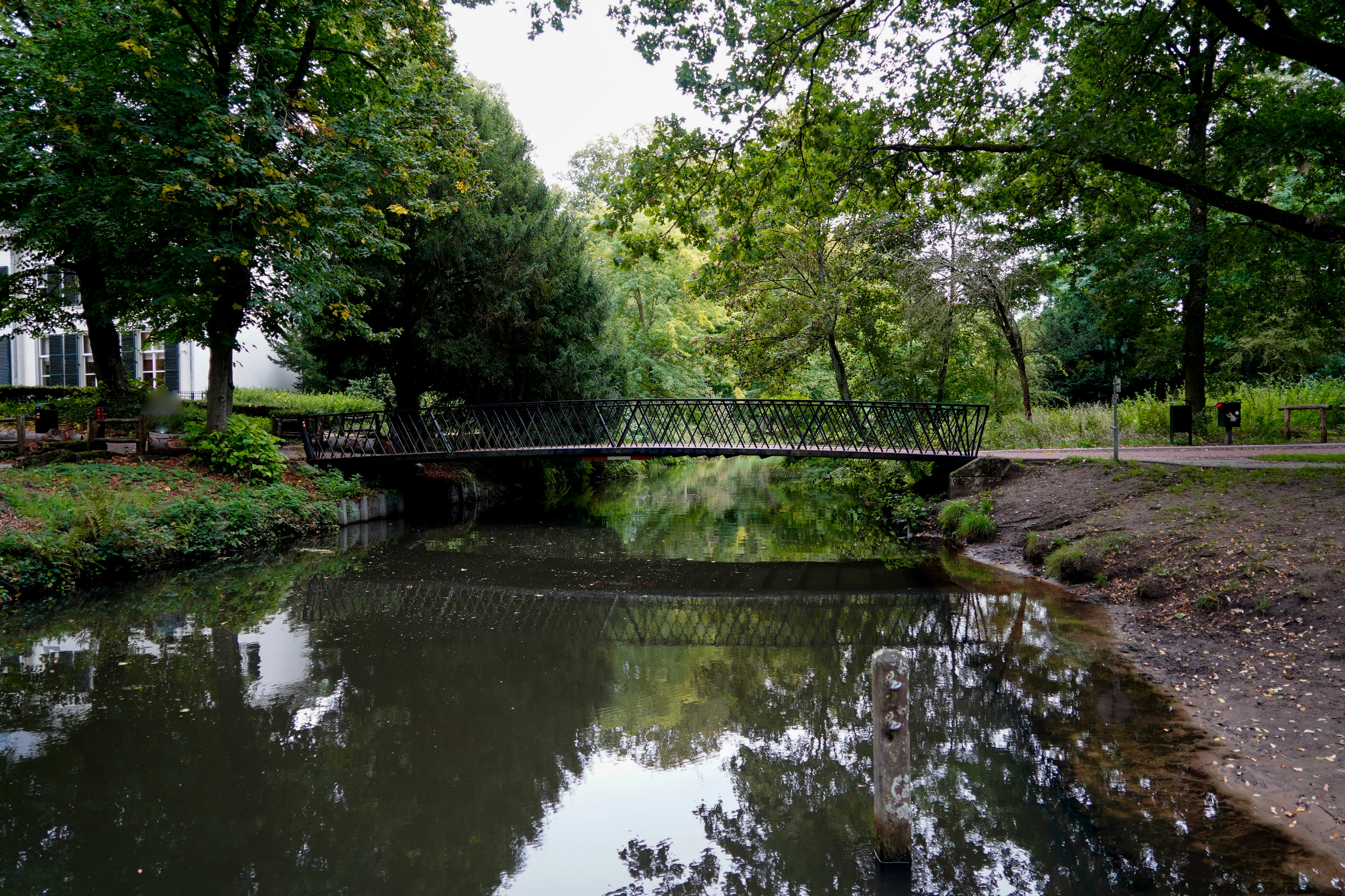 Eine Metall-Fußgängerbrücke, die einen ruhigen Kanal überquert, umgeben von üppig grünen Bäumen