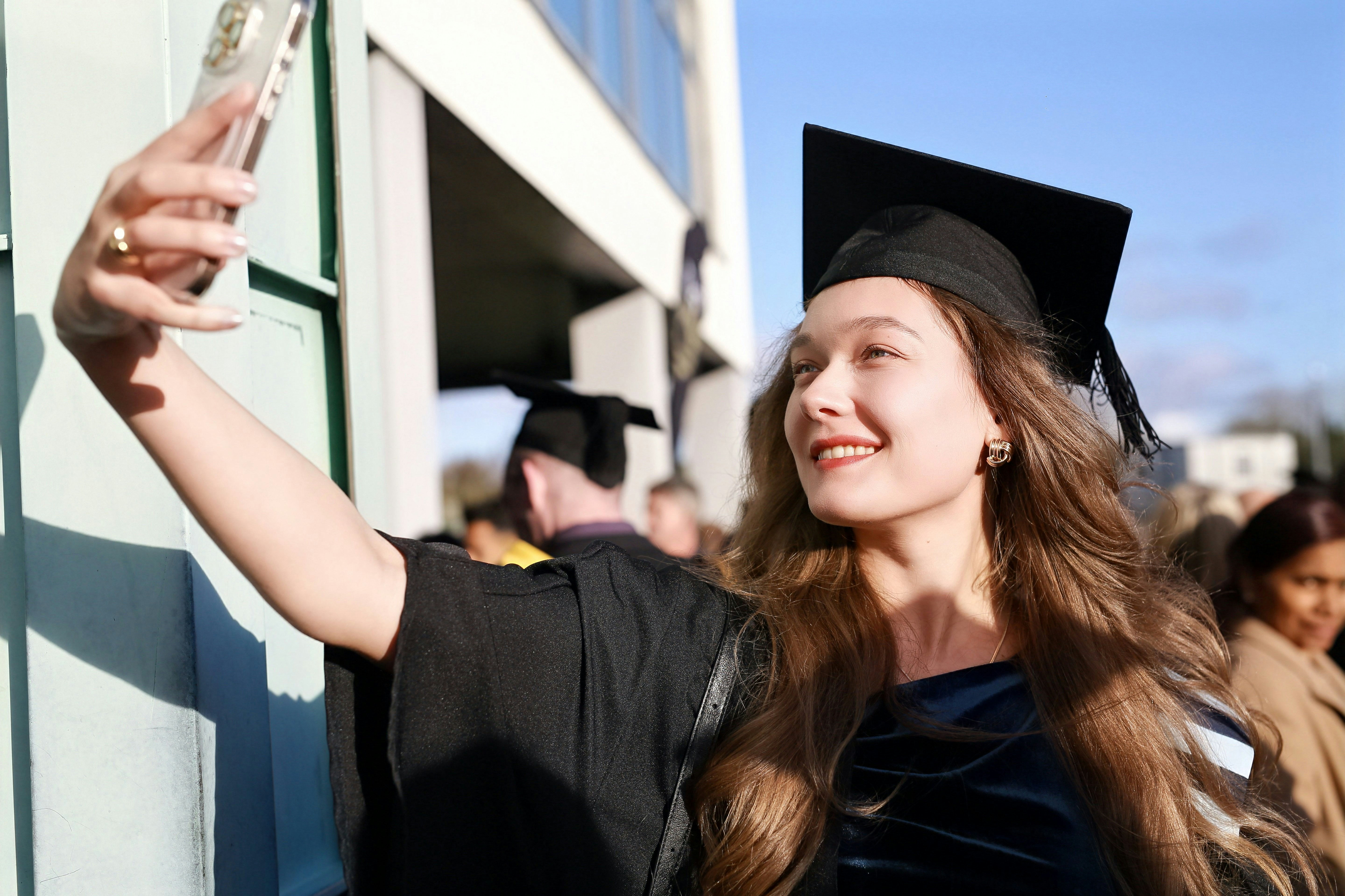 A smiling woman in a graduation cap and gown taking a selfie outdoors
