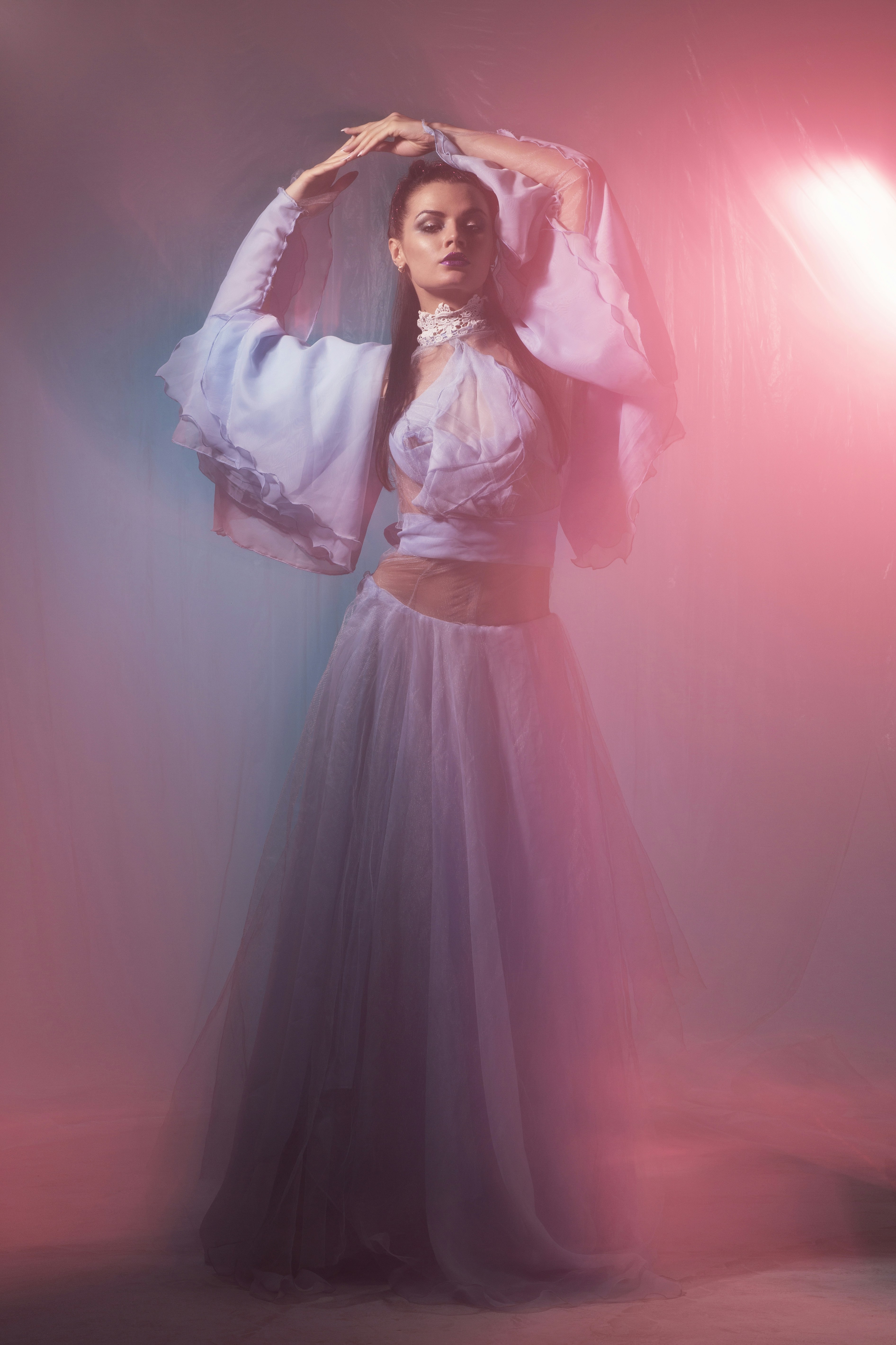 A woman in a flowing light blue dress with arms raised under pink light