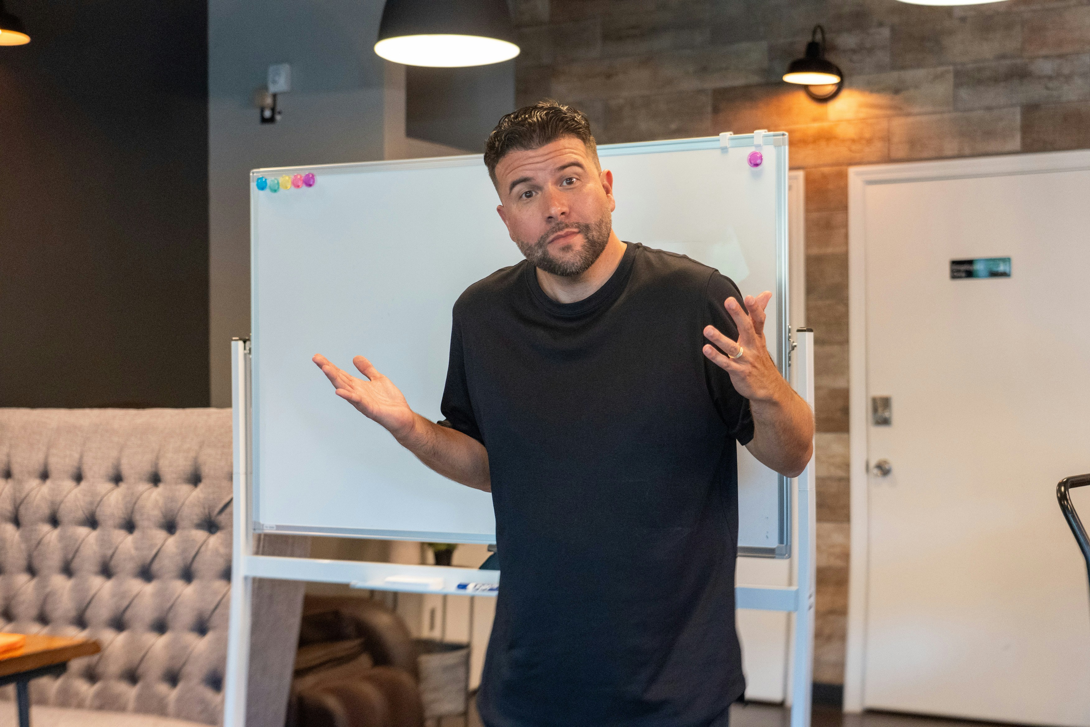 A man in a black t-shirt gesturing with open hands before a whiteboard