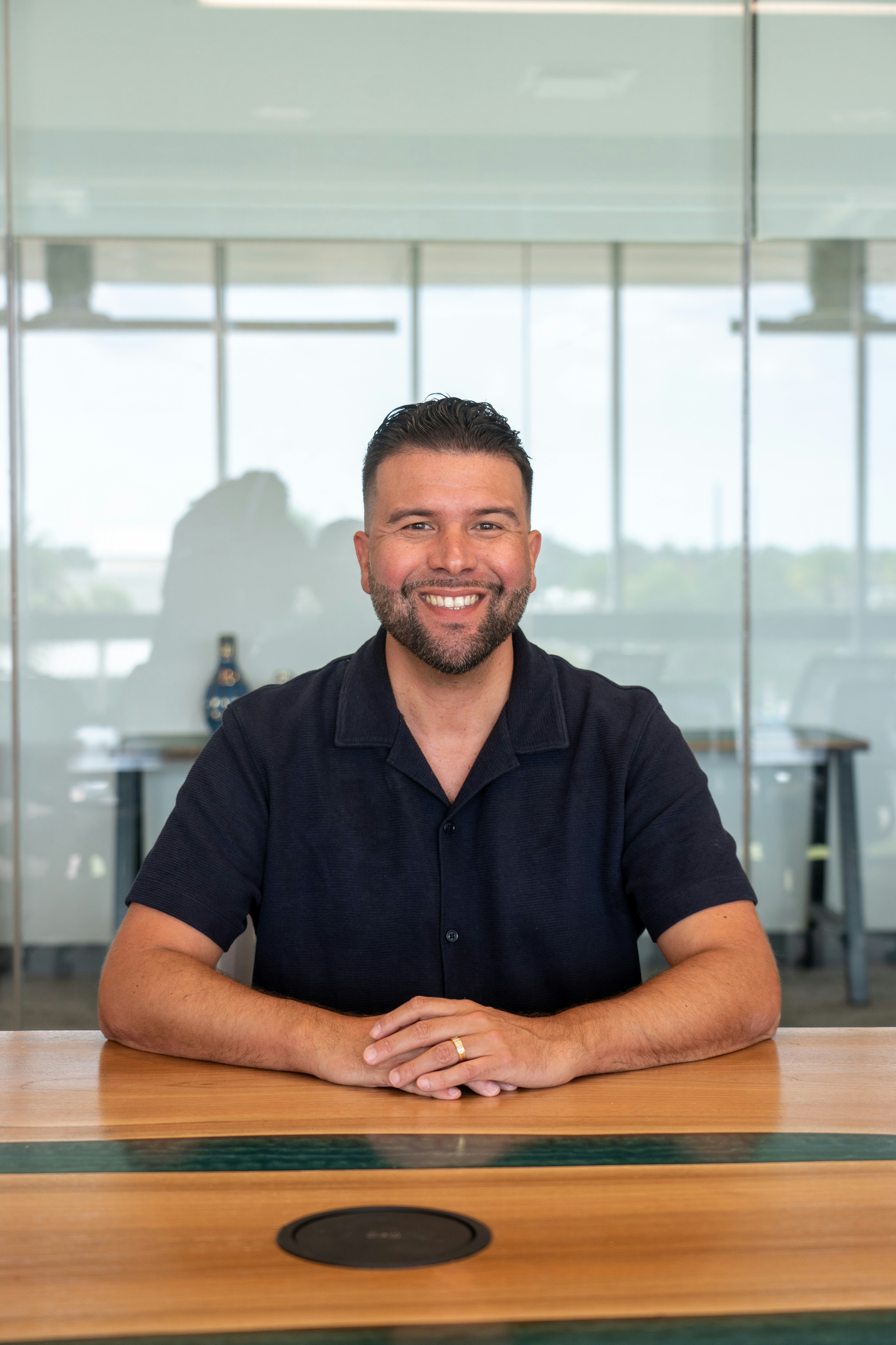 A smiling man in a dark polo shirt sitting at a wooden table