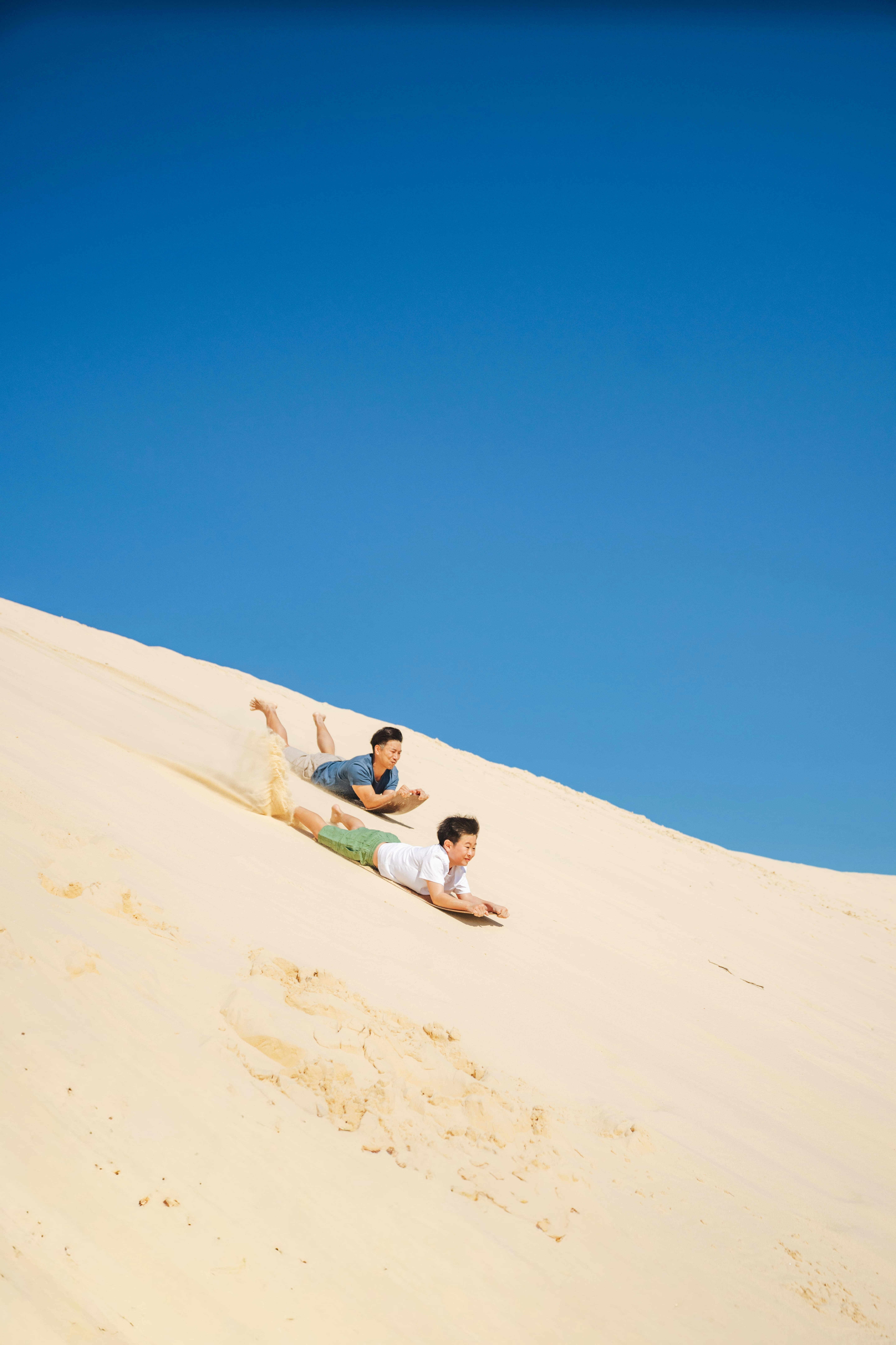 Two people sandboarding down a steep sand dune under a clear blue sky