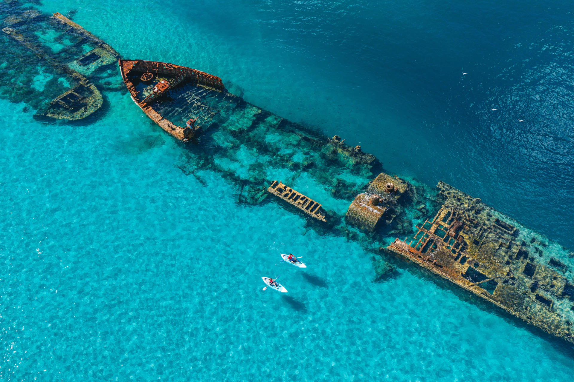 Two people kayaking near a large rusted shipwreck in clear turquoise water