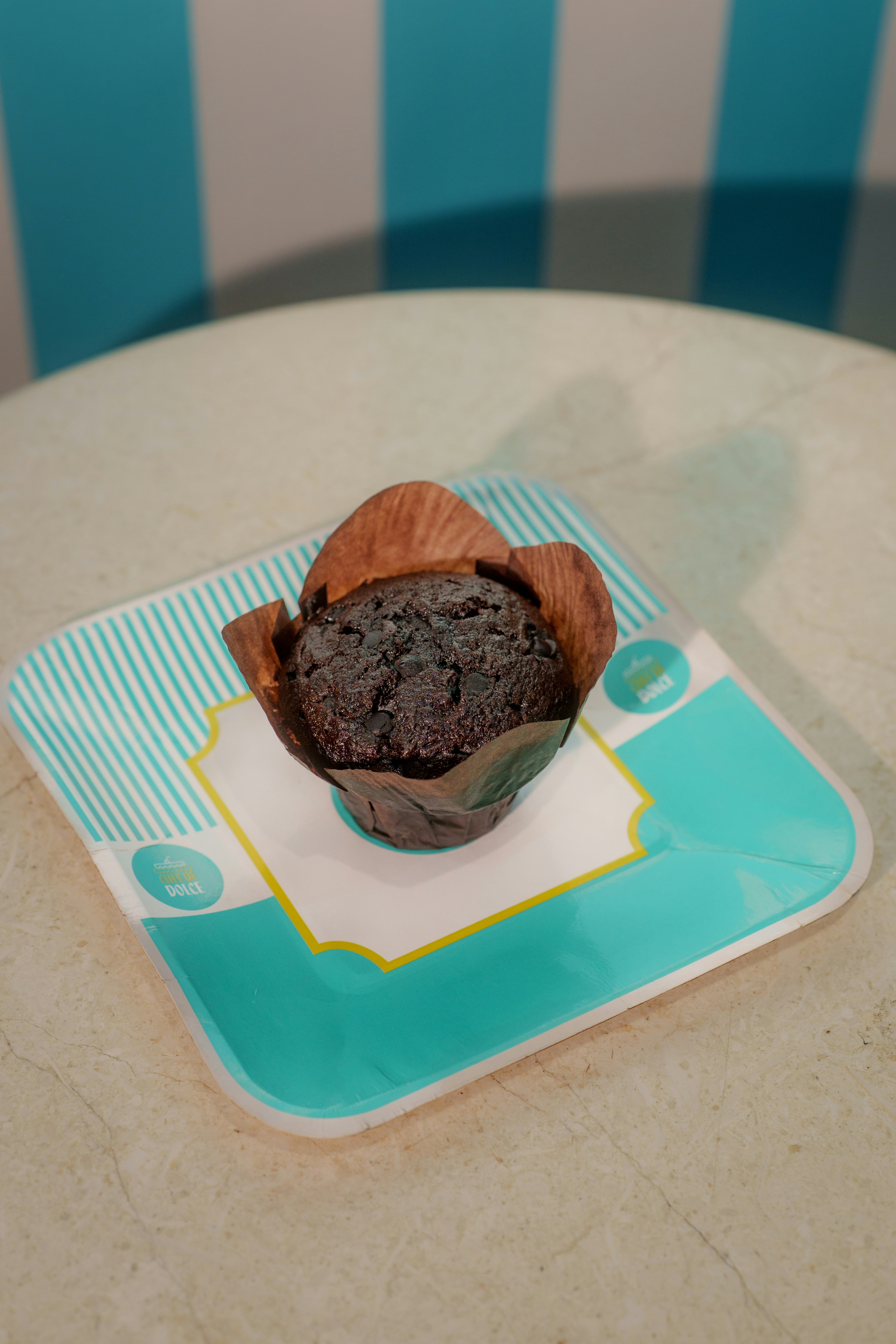 A chocolate chip muffin in a brown wrapper on a paper plate
