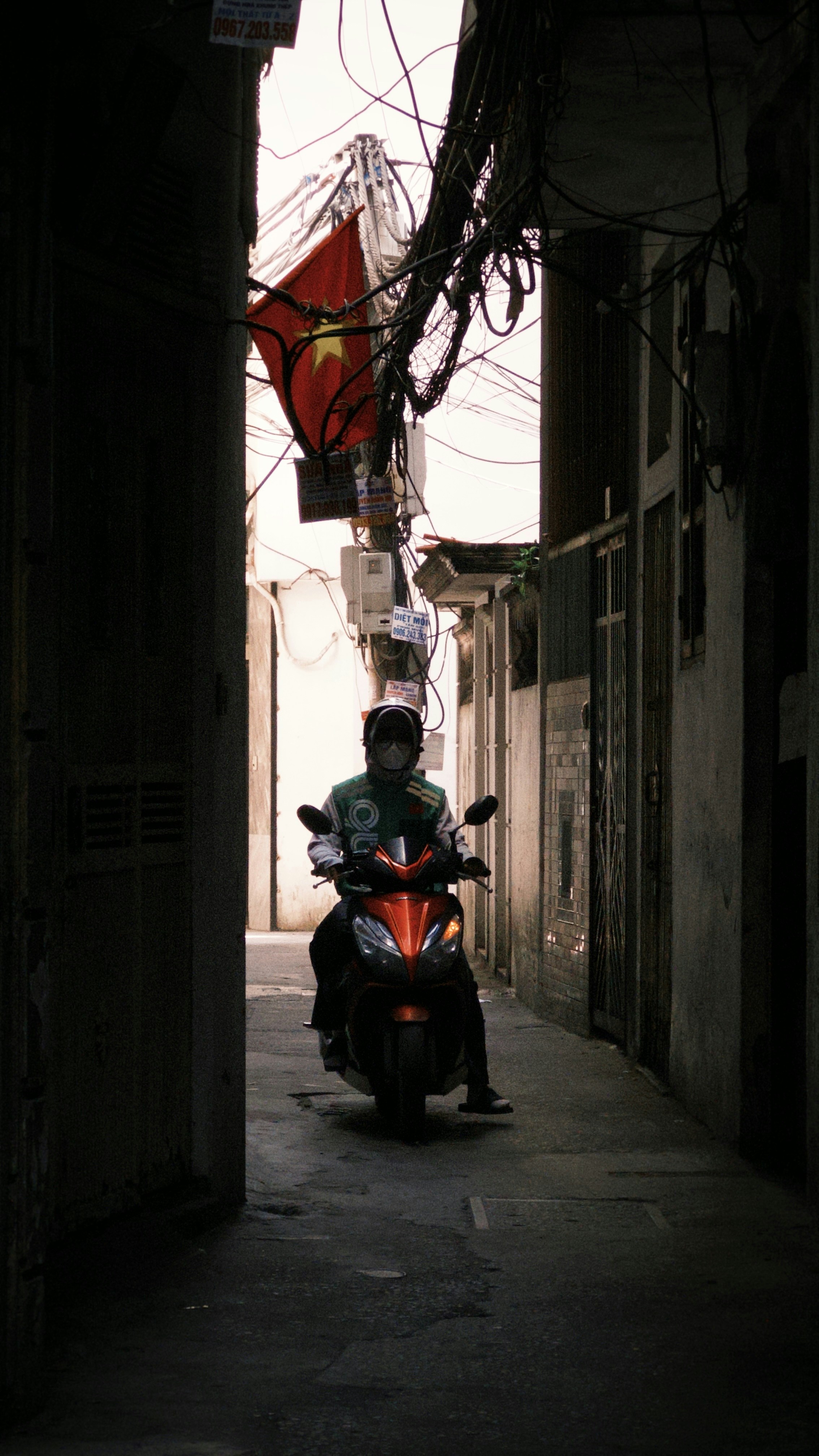 A person on an orange scooter in a narrow alley under tangled wires
