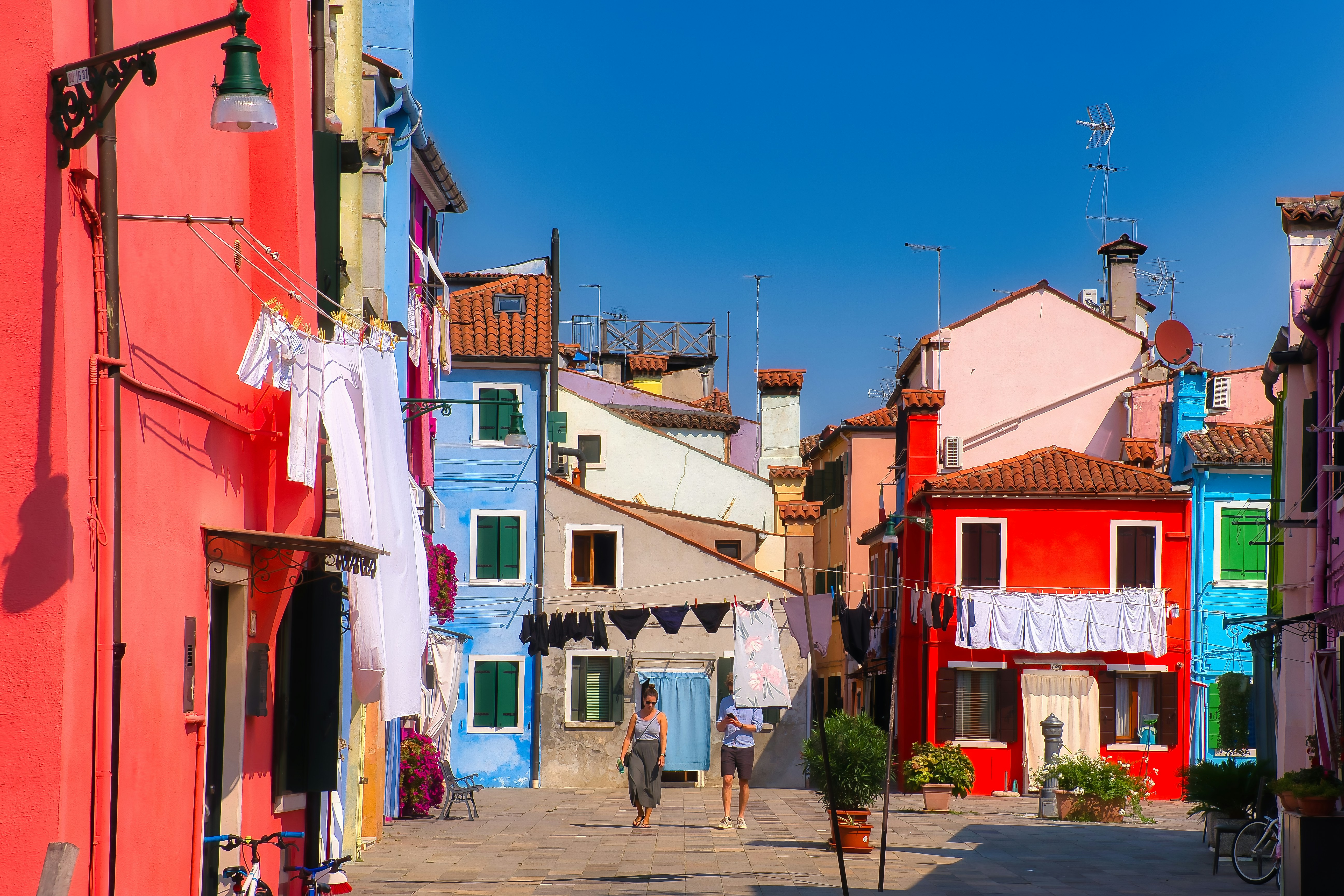 Two people walking through a sunny courtyard between colorful houses with hanging laundry