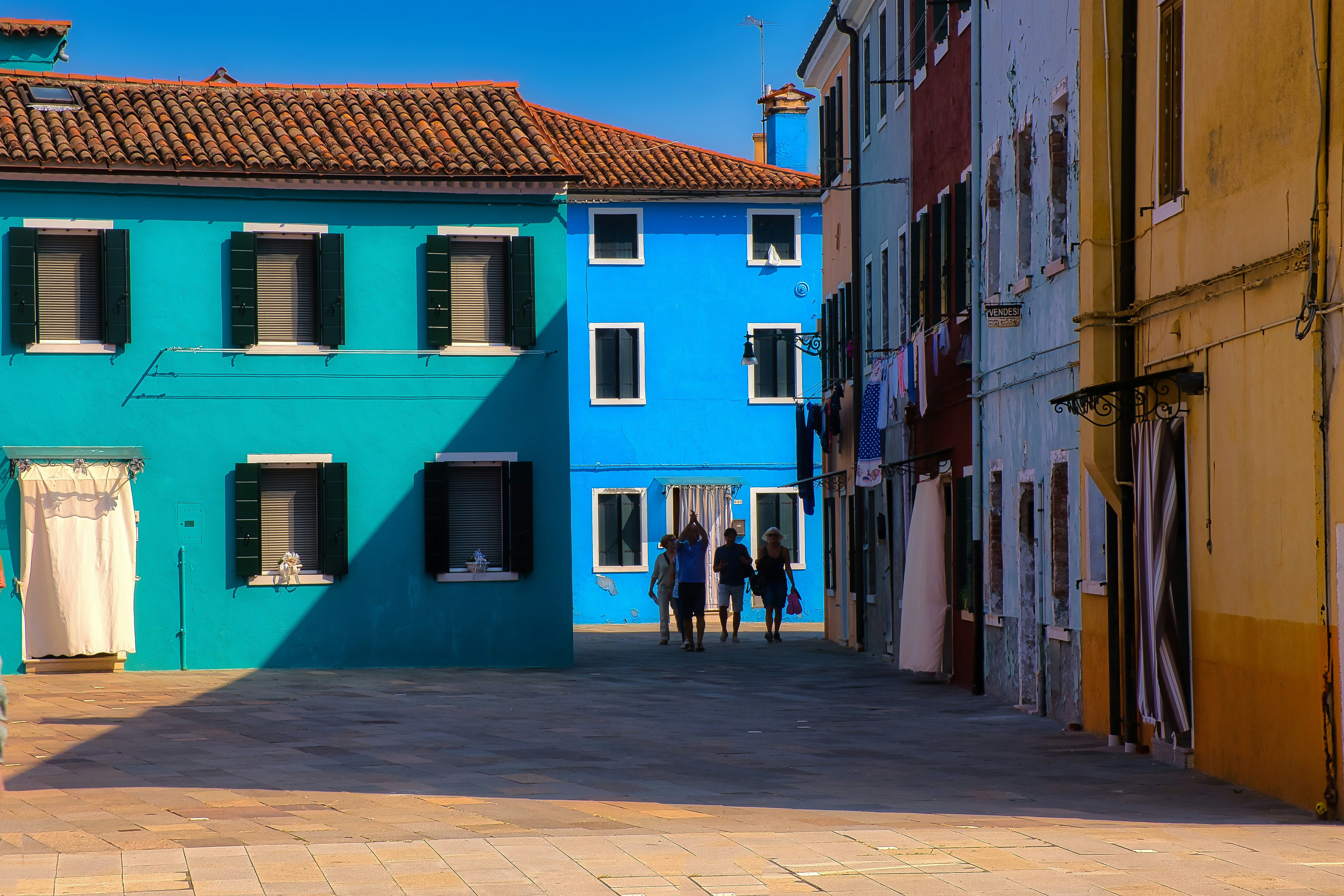A group of people walking past brightly painted houses in a sunlit square