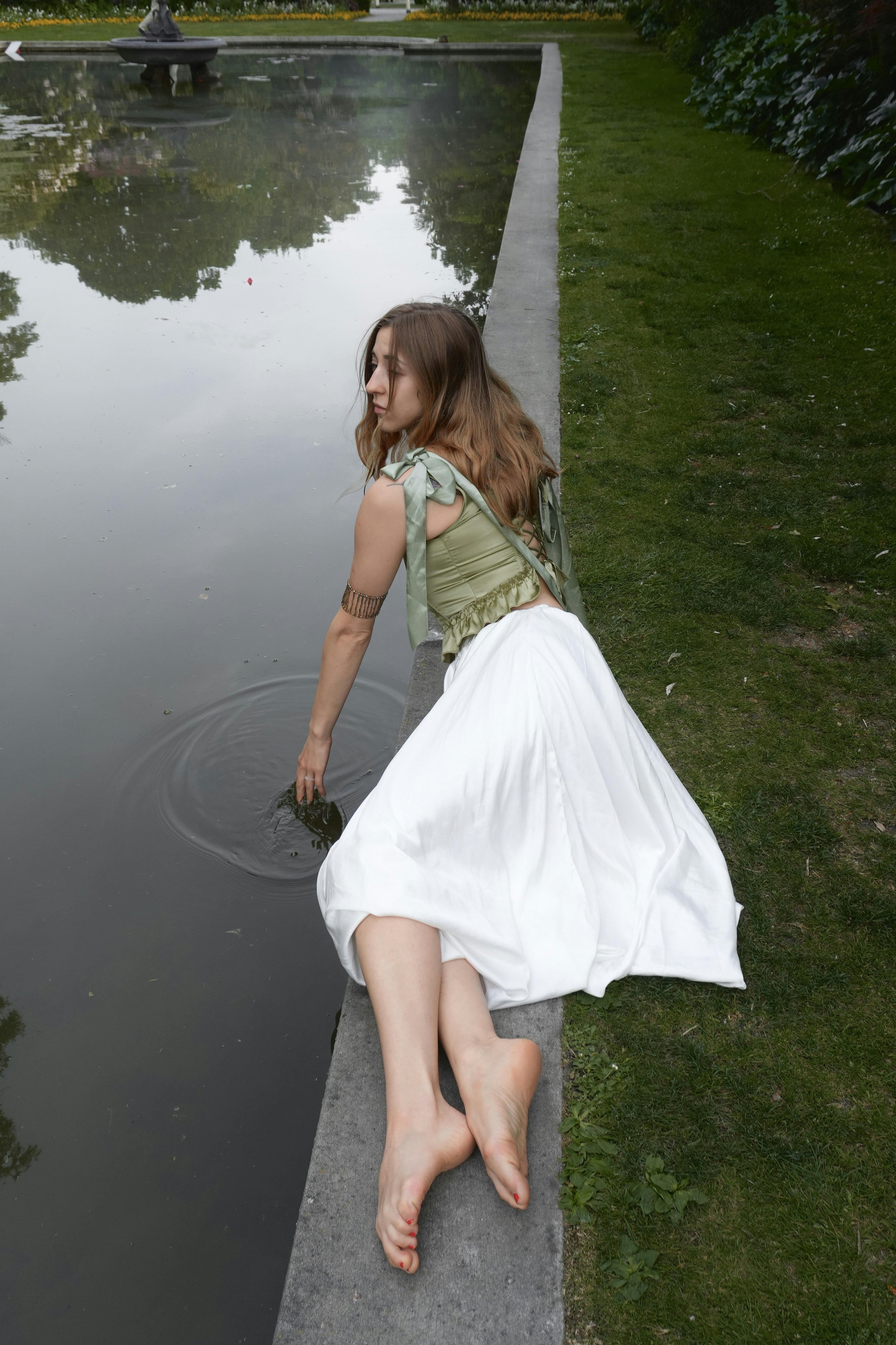 A woman in a green corset and white skirt touching a pond surface